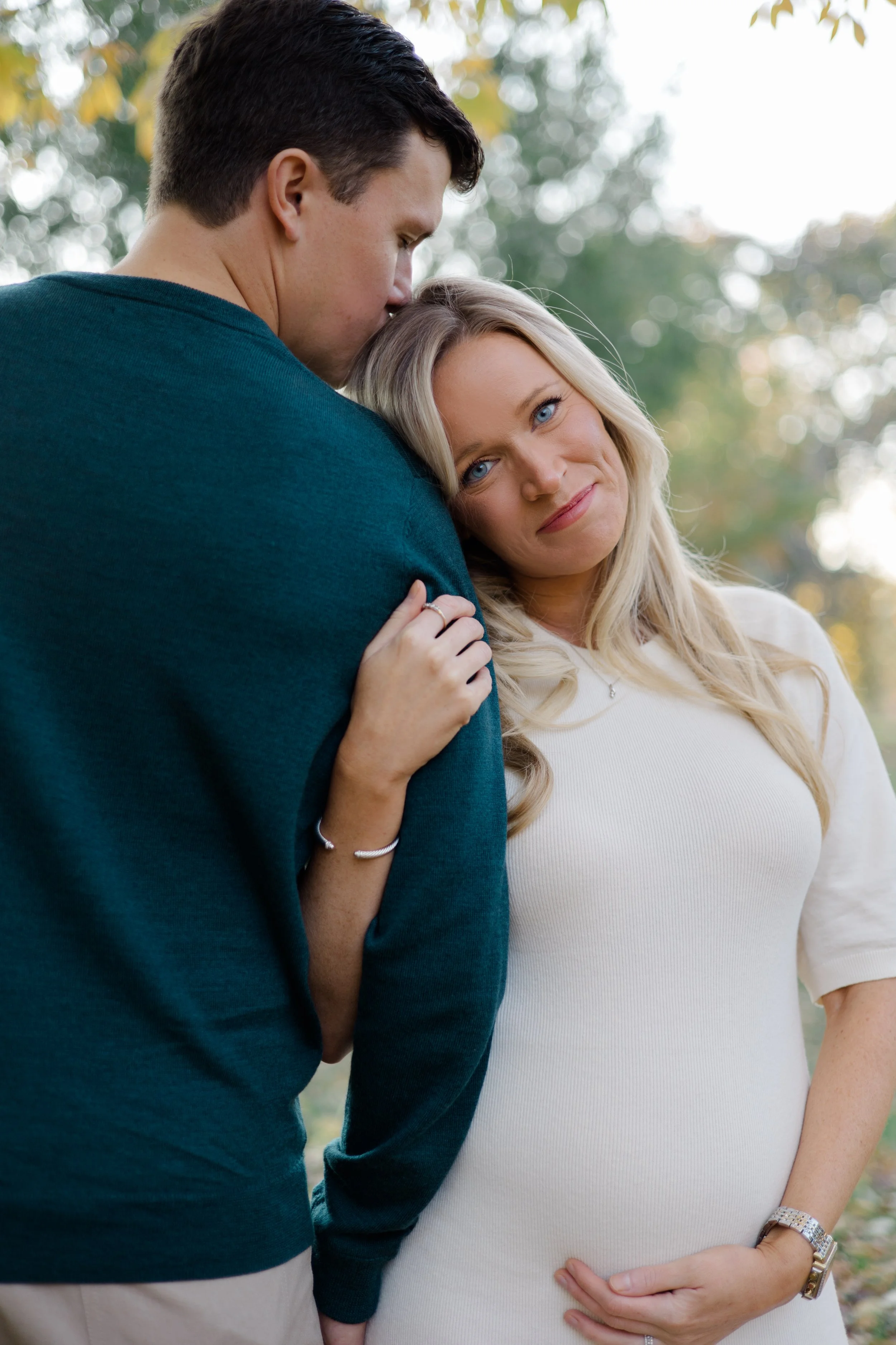 A man and a pregnant woman outdoors, with the man kissing the woman's forehead, and the woman gently holding her pregnant belly, smiling softly.