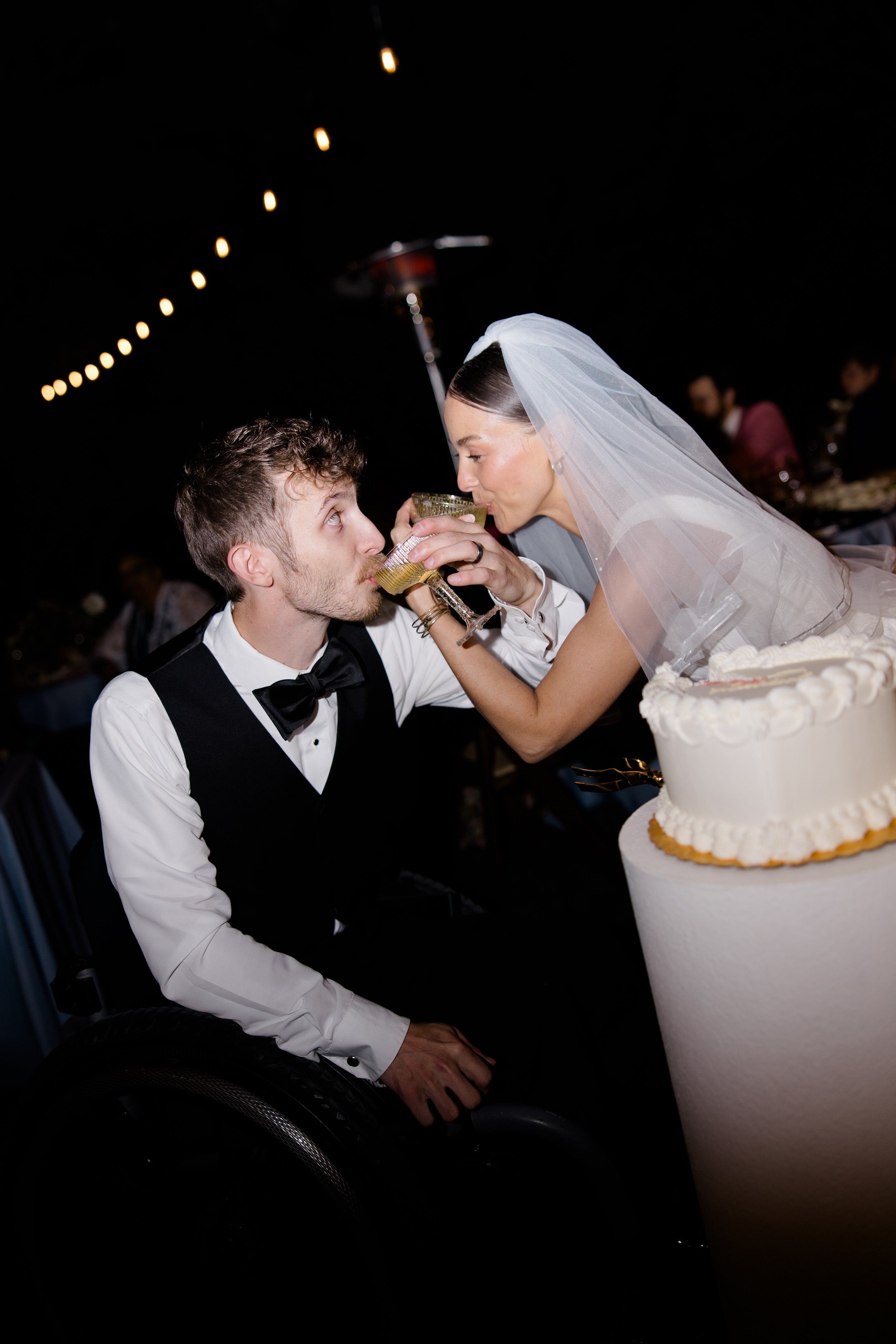 A woman in a wedding dress and veil shares a drink with a man in a tuxedo at a wedding reception. There is a wedding cake in front of them, and a dark background with string lights overhead.