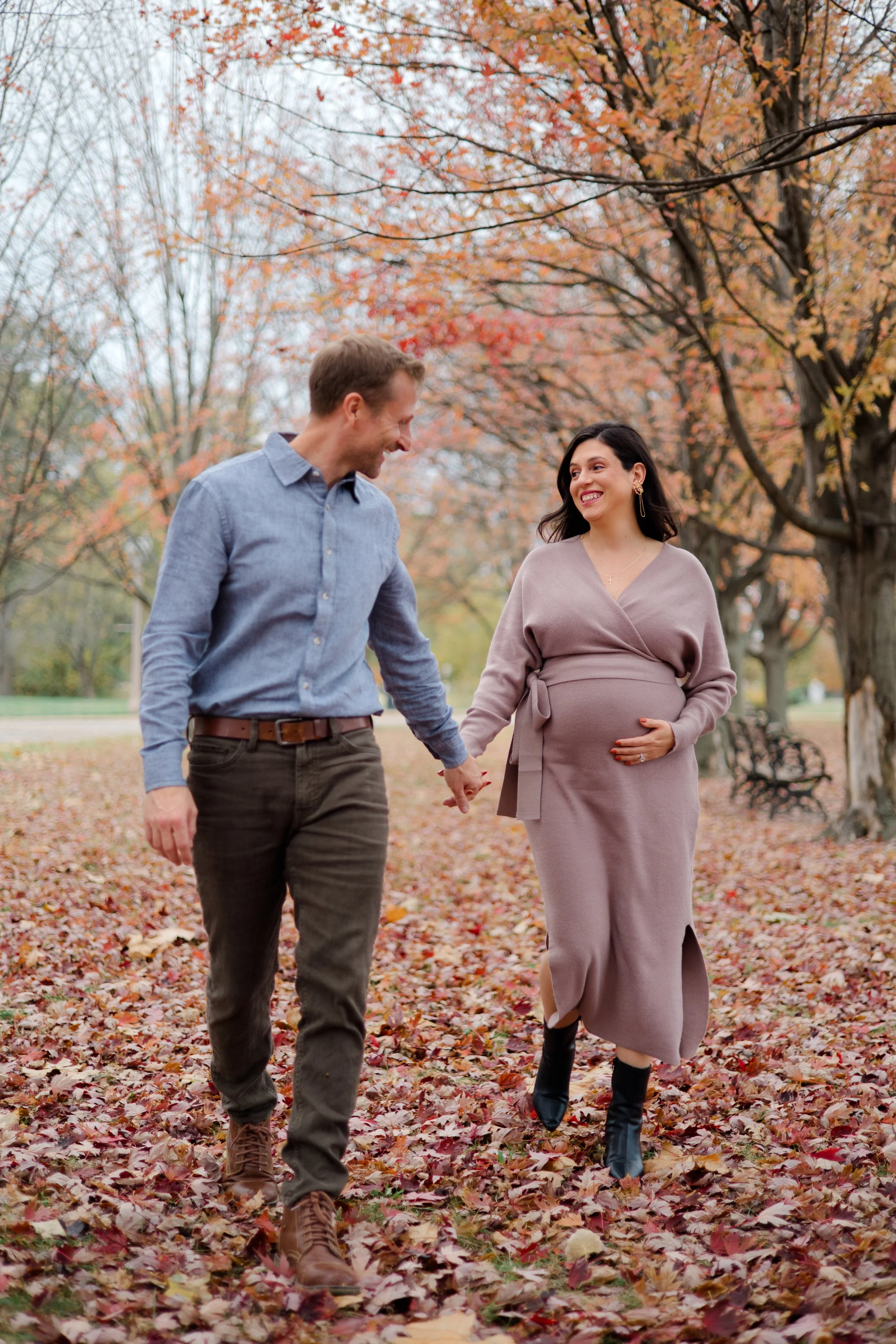 A couple walking hand in hand through a park covered in fallen autumn leaves, with trees displaying orange and yellow foliage in the background. The woman is pregnant, wearing a light mauve dress and black boots, smiling at the man; the man is wearin