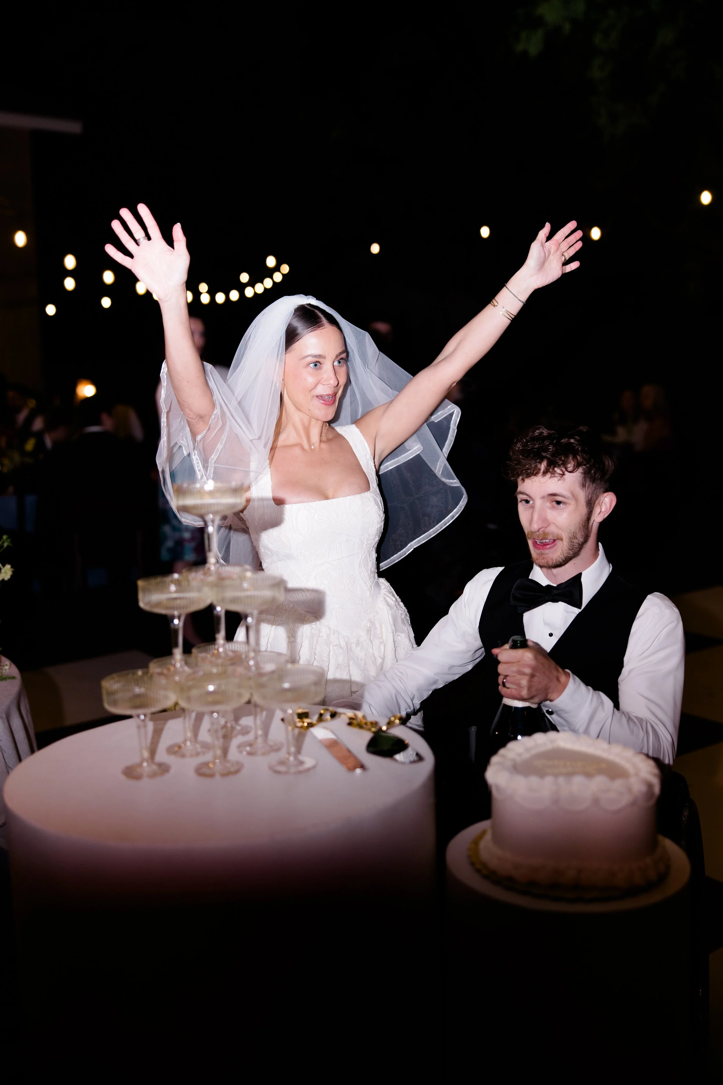 A woman wearing a wedding dress and veil with her arms raised, celebrating at her wedding reception. Beside her is a man in a tuxedo and bow tie, sitting at a table with a domino-shaped champagne tower, a wedding cake, and other decorations. There ar