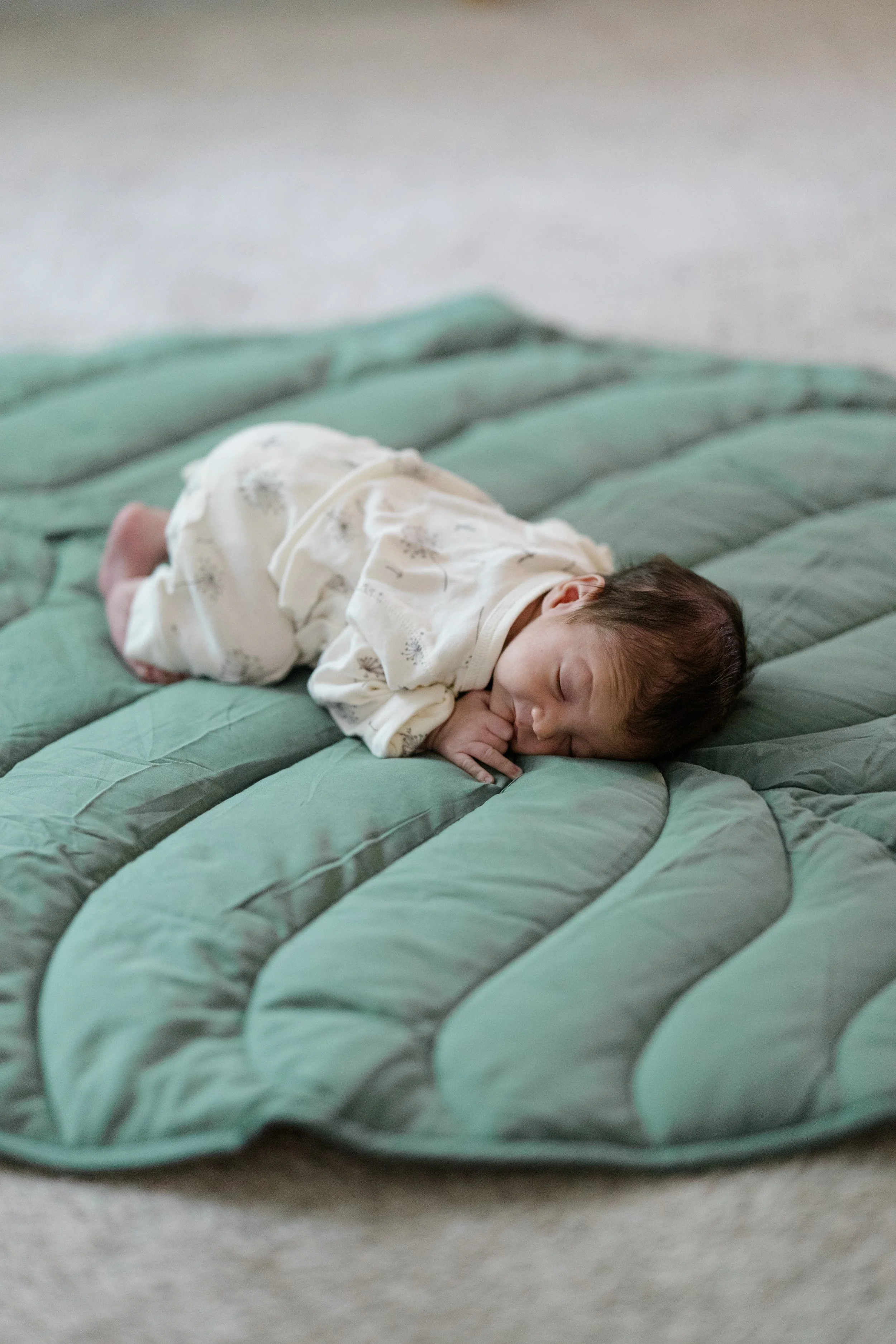 A newborn baby lying on a green quilted bedspread, wearing a white onesie with light-colored patterns, sleeping peacefully with one hand near his face.