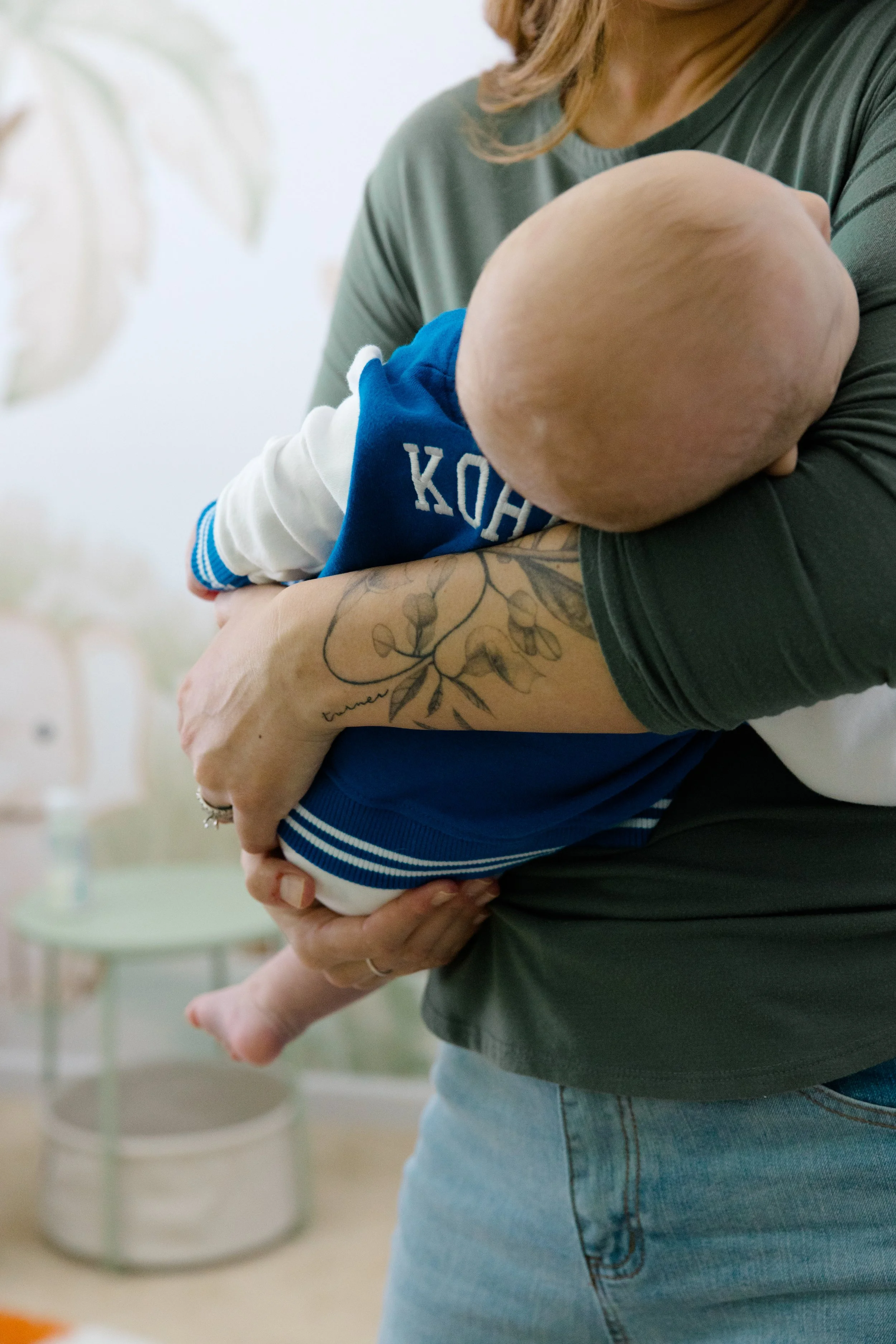 A woman holding a baby in her arms indoors. The baby is wearing a blue and white jacket with the word 'KOH' visible. The woman has a tattoo on her forearm and is wearing a dark green shirt and light blue jeans.