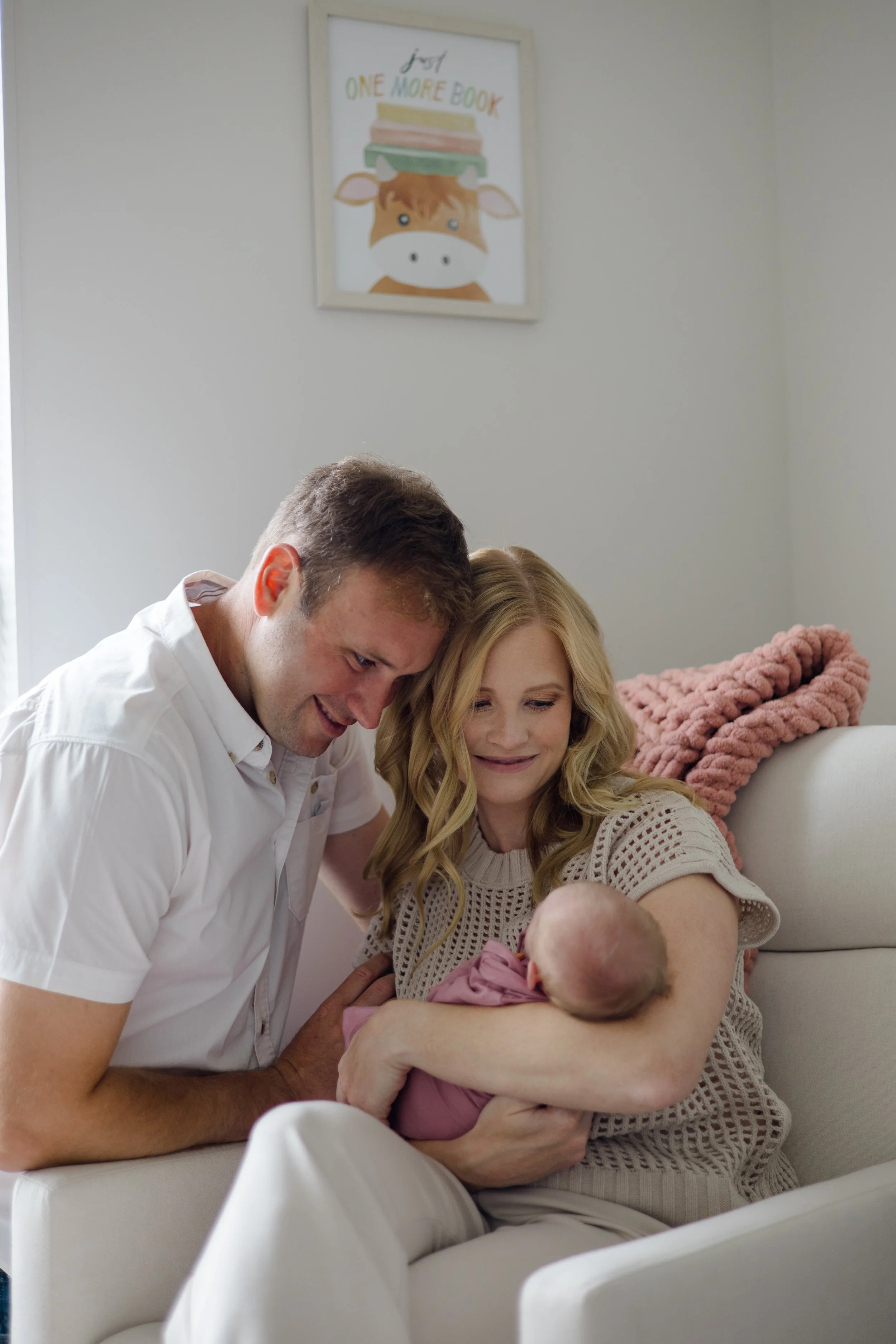 A woman holding a baby with a man leaning over her, all smiling, in a cozy living room with a framed picture of a cow and a pile of books on the wall behind them.