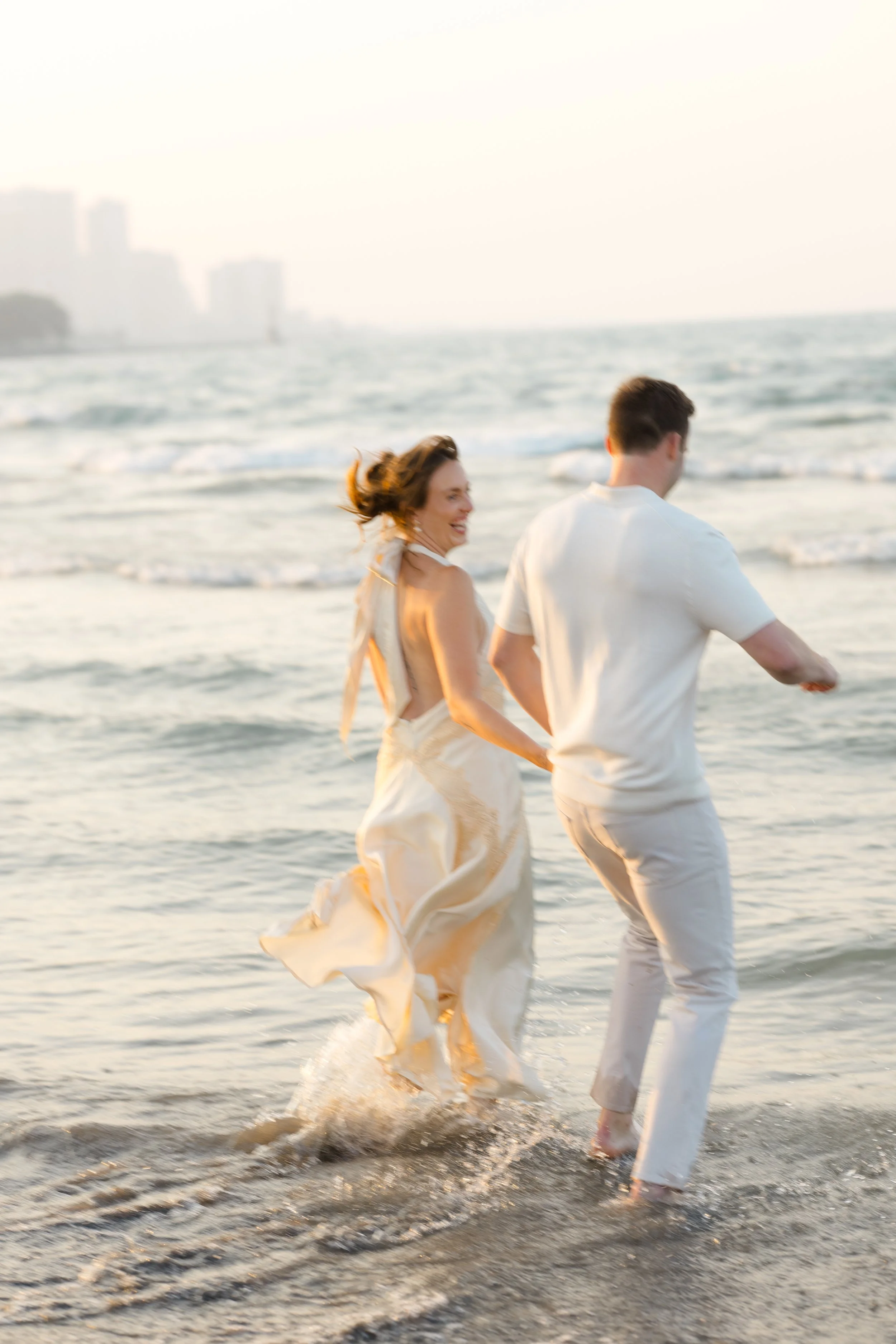 A couple walking hand in hand along the shoreline at the beach during sunset, with waves gently crashing at their feet.