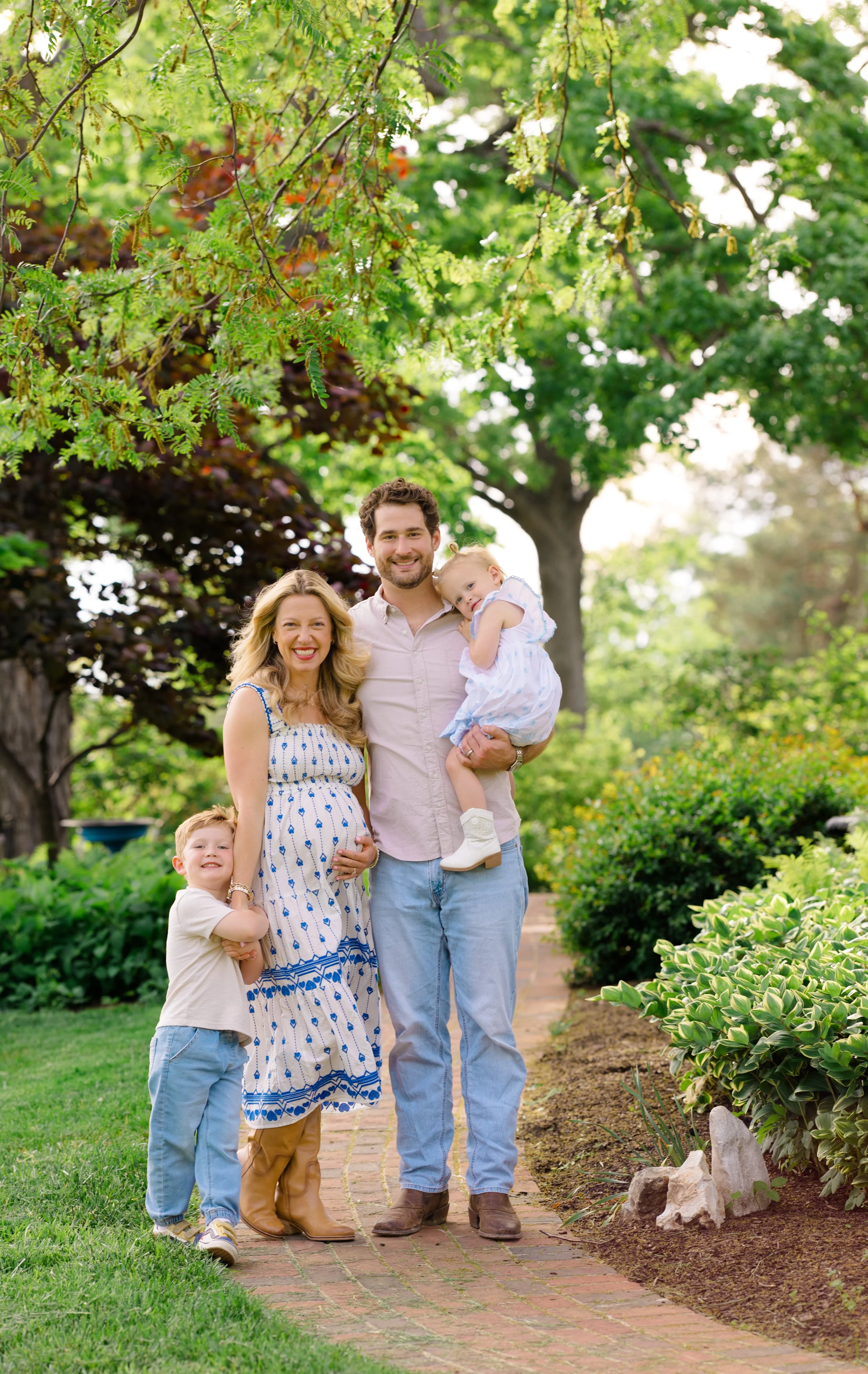 A family of five posing and smiling outdoors on a brick pathway surrounded by green trees and plants.