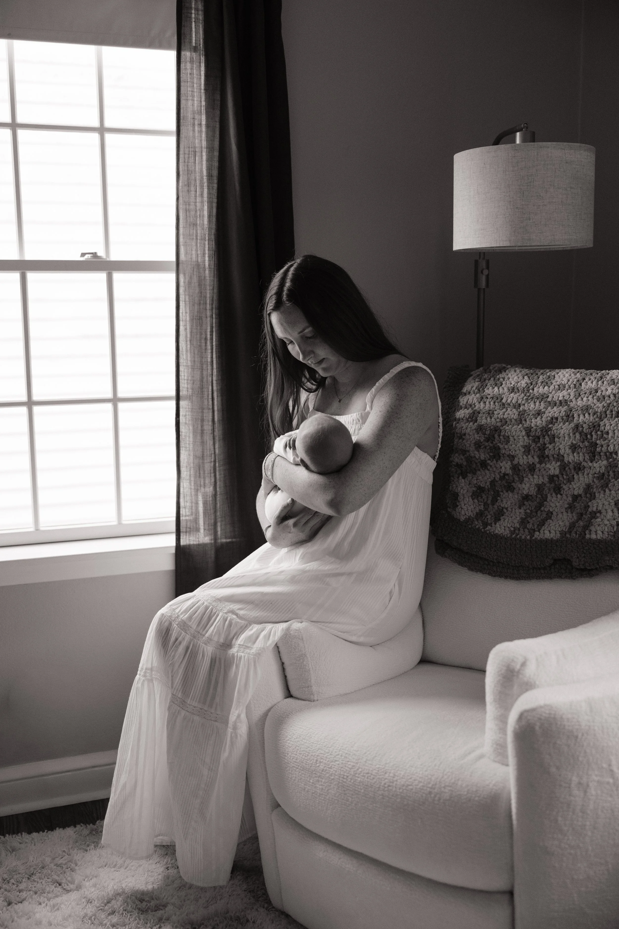 A woman in a white dress holding a baby while sitting on a cushioned chair in a room with a window and a lamp.