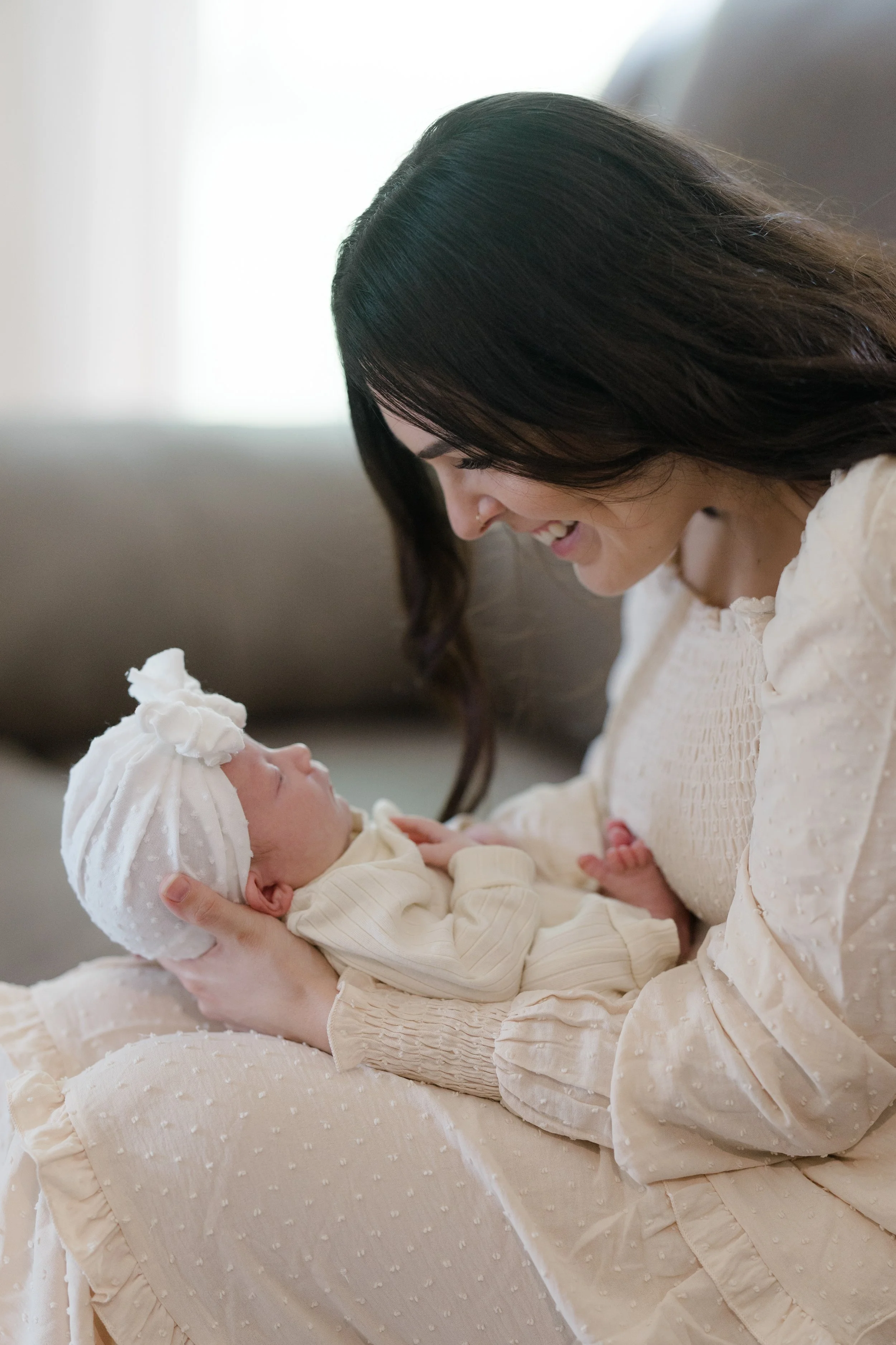 A woman with dark hair smiling and looking down at a newborn baby she is holding. The baby is wearing a white knit hat and cream-colored clothing, and is resting comfortably in the woman's arms.