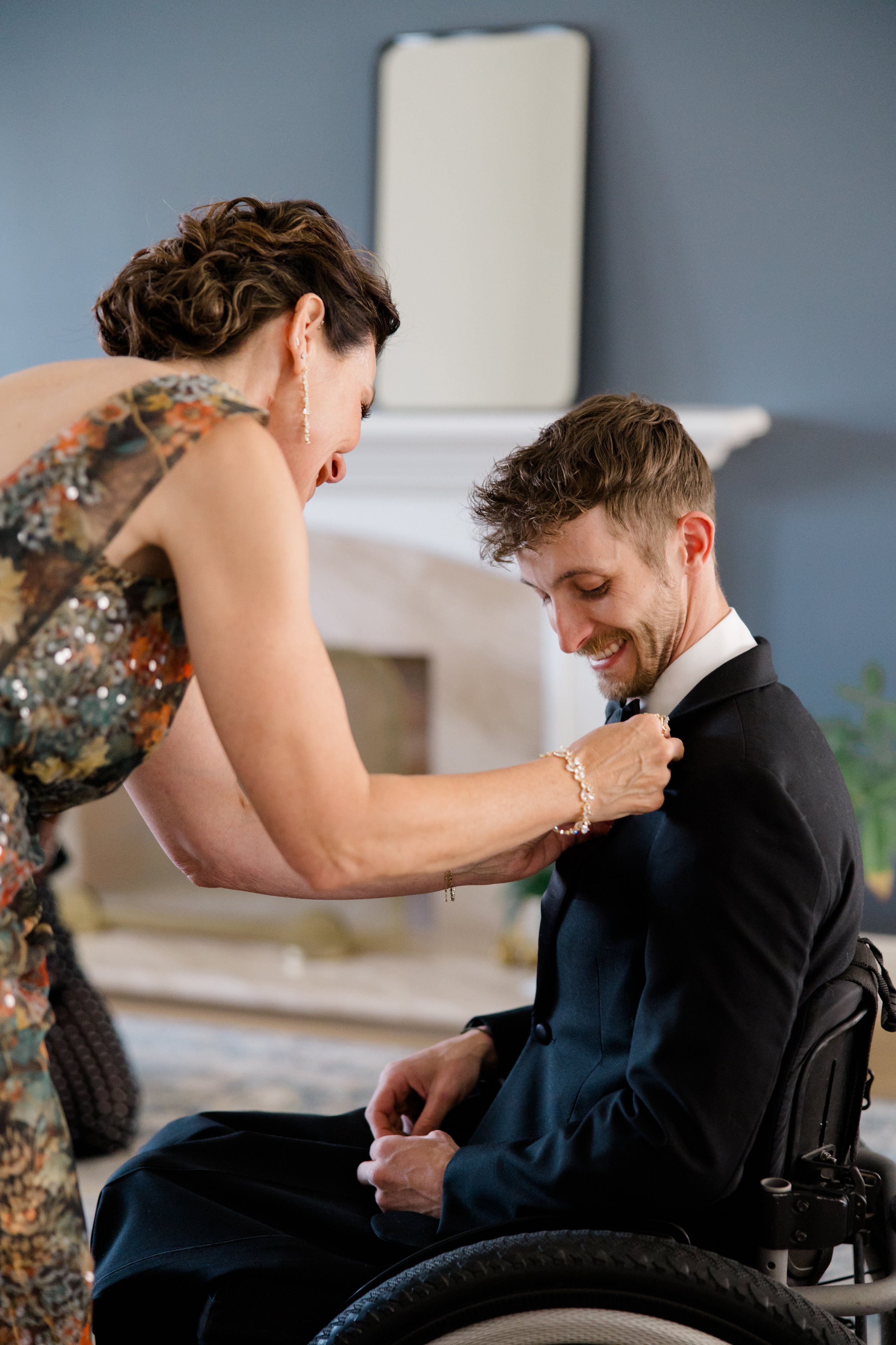 A woman in a floral dress pins a corsage on a man in a wheelchair dressed in a tuxedo.