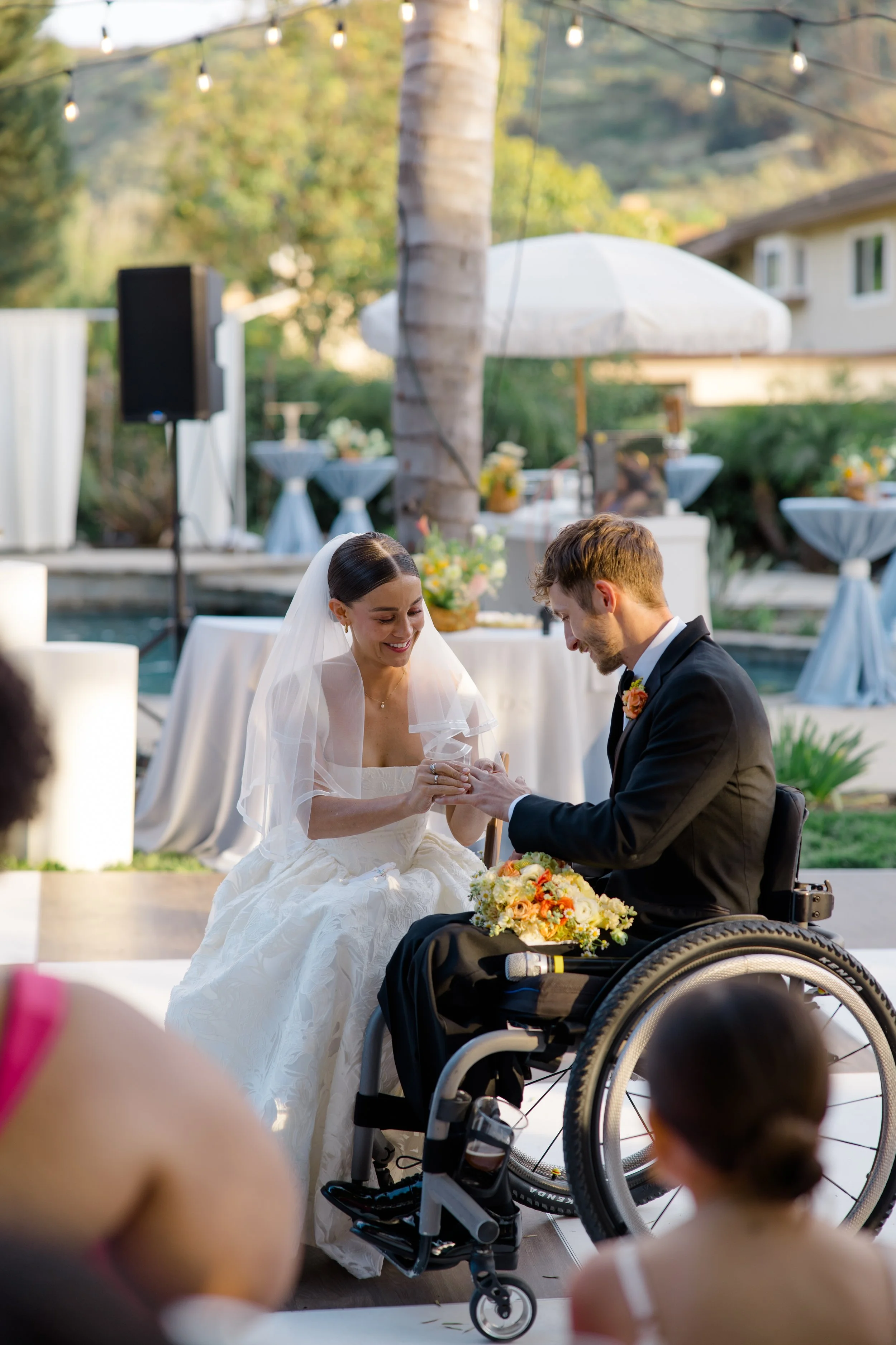 A bride and groom exchange vows during an outdoor wedding ceremony. The bride is sitting in a wedding dress and veil, while the groom is in a suit and a wheelchair. The background shows decorated tables, an umbrella, and string lights, with trees and