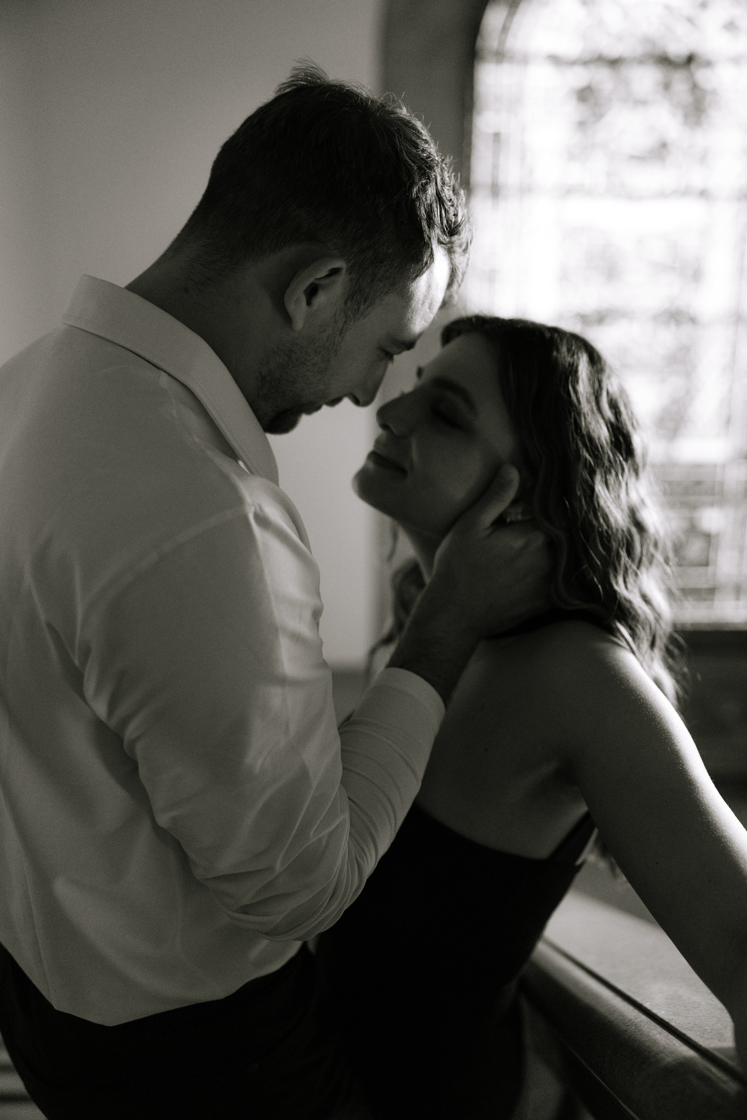 A black and white photograph of a man and a woman engaging in an intimate moment, holding each other's faces close with their foreheads touching, in a softly lit room with a window in the background.