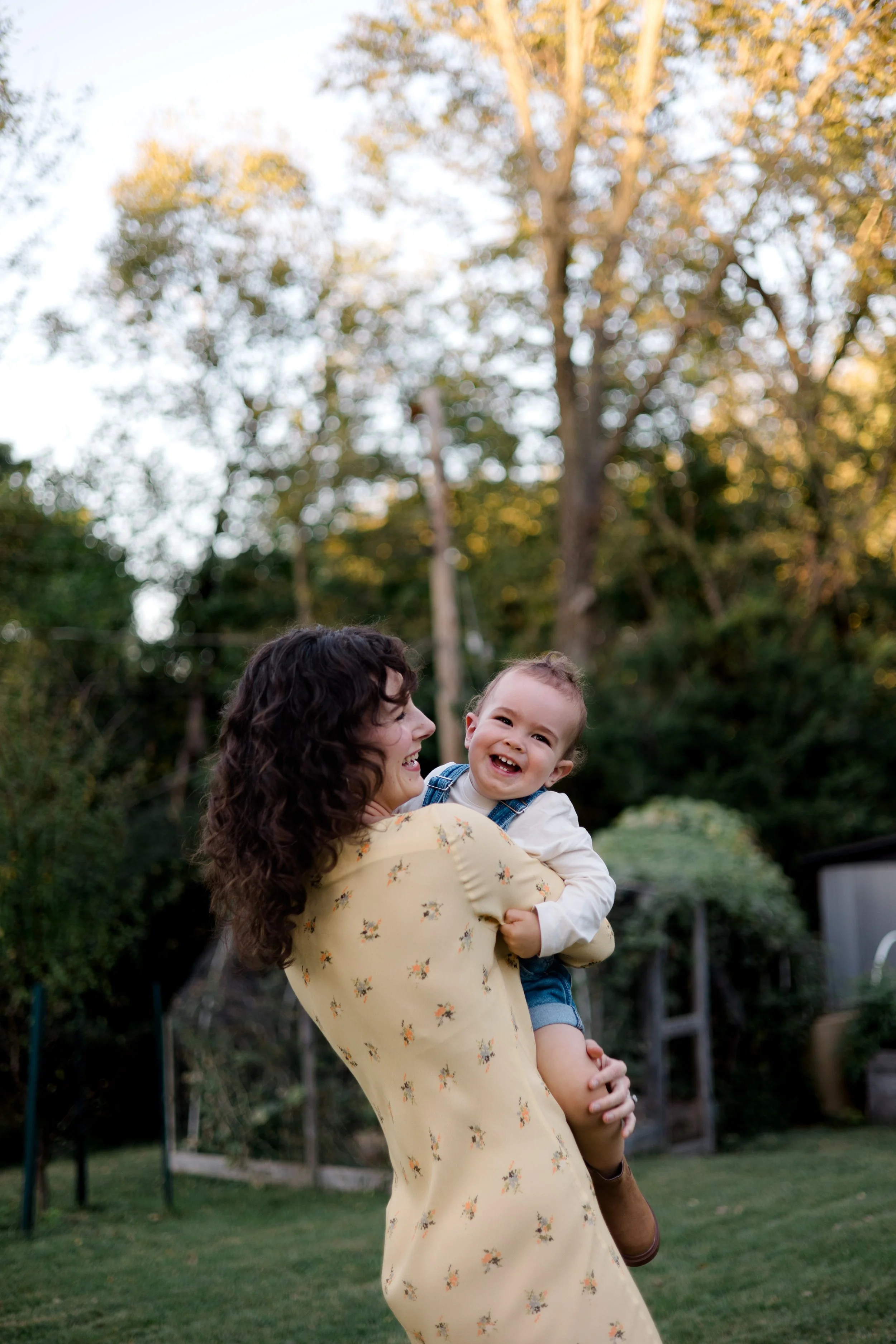 A woman with curly brown hair holding a smiling young boy outdoors in a backyard with trees and grass, during the evening.