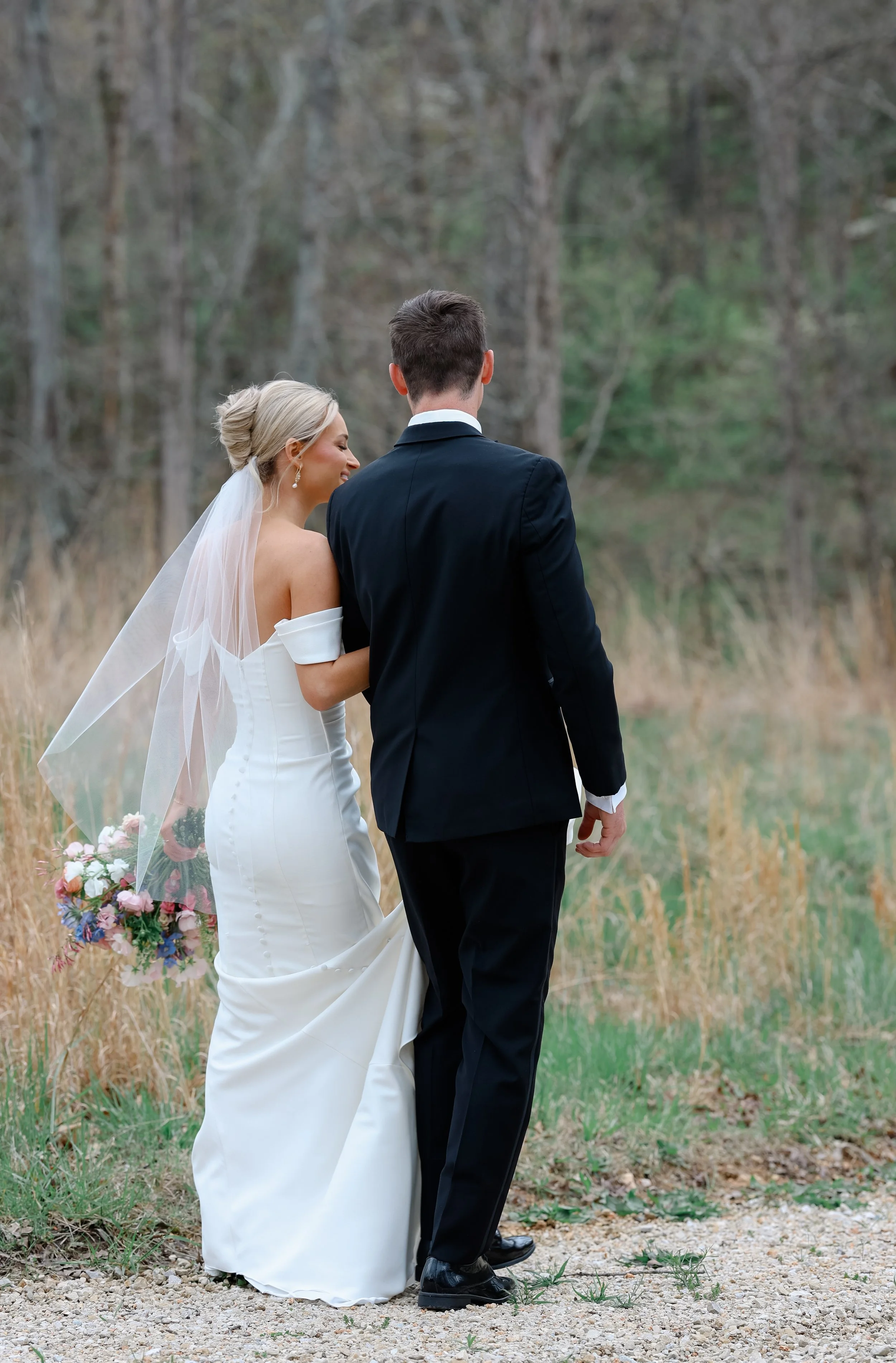 A bride and groom walk outdoors in a natural setting with trees and grass, holding hands and smiling at each other. The bride is wearing a white wedding dress with off-the-shoulder sleeves and a veil, carrying a bouquet of flowers. The groom is dress