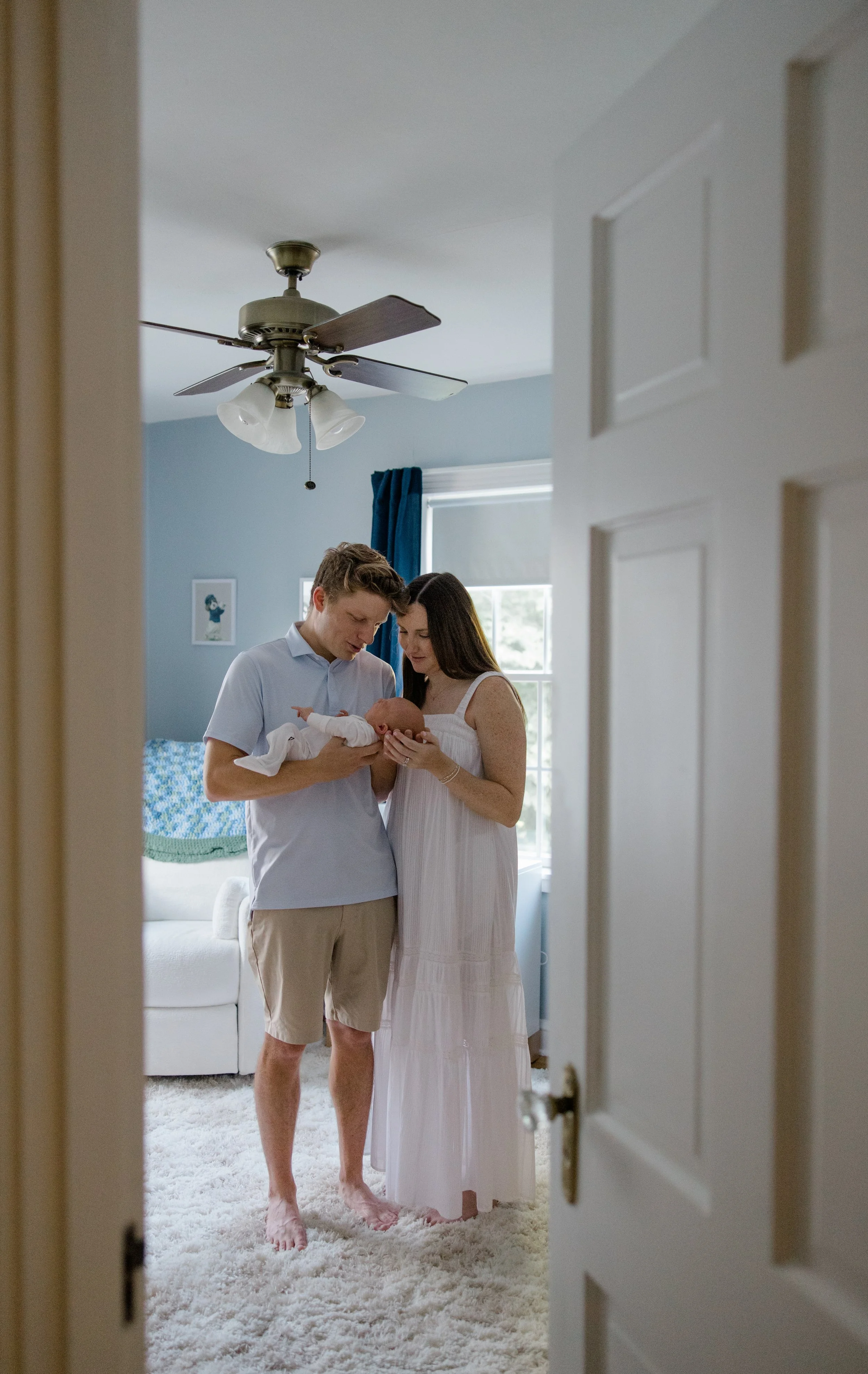 A couple holding a newborn baby in a bedroom, seen through a partially open door, with a ceiling fan and window in the background.