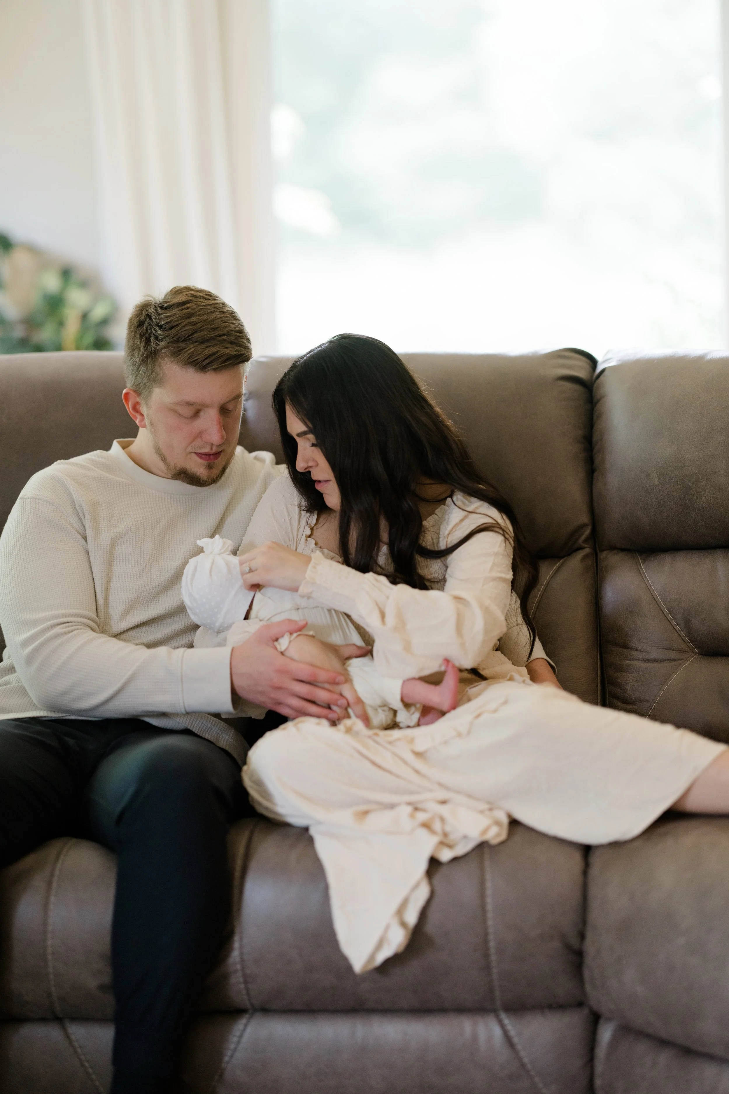 A man and woman sitting on a brown couch in a living room, holding a newborn baby.