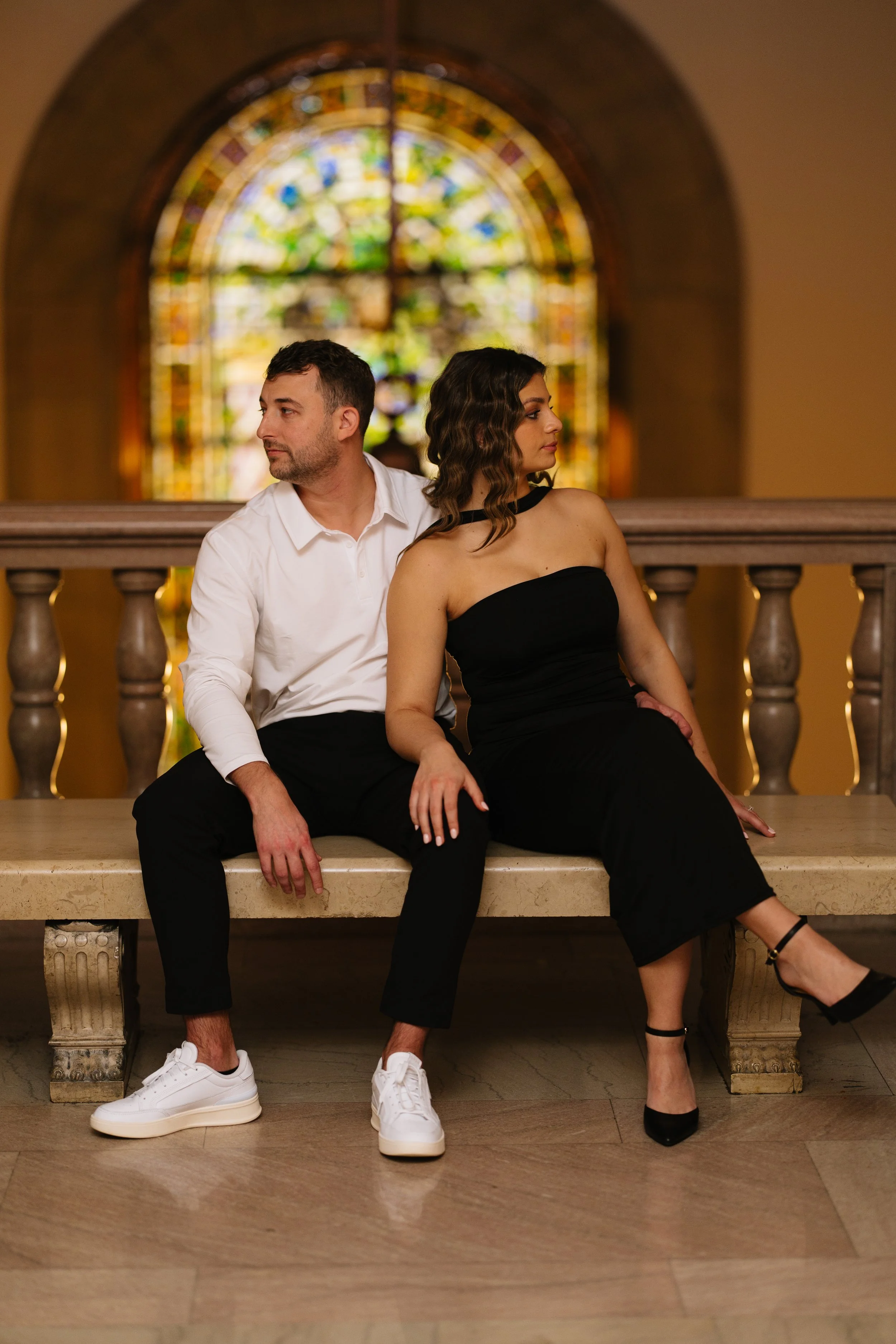 A man and a woman sitting on a bench inside a church with a stained glass window in the background.