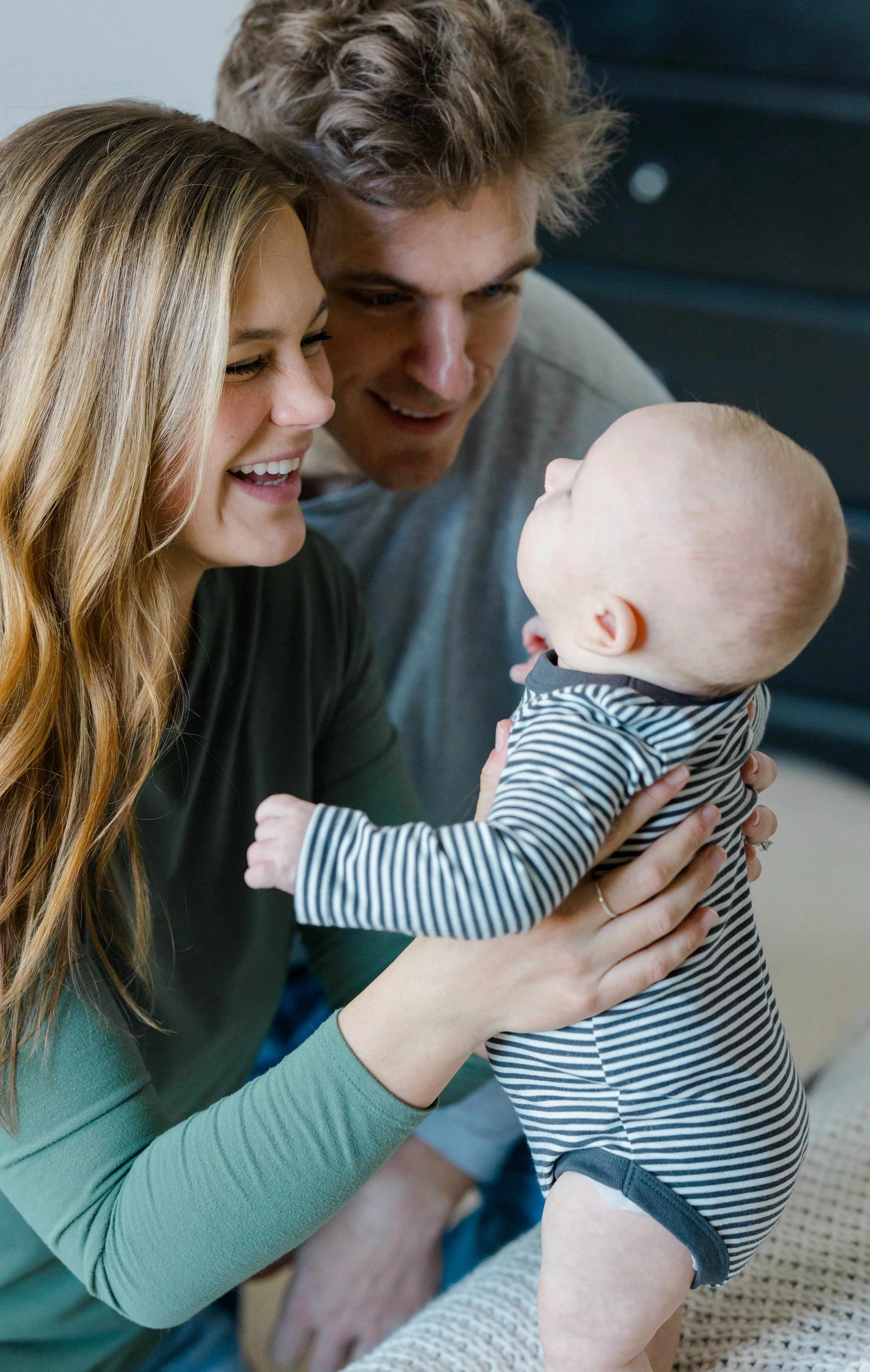 A young woman and a young man smiling and looking at a baby. The woman is wearing a green long sleeve shirt, and the man is behind her with a beige shirt. The baby has a bald head, is wearing a striped outfit, and is facing the woman and man, who are