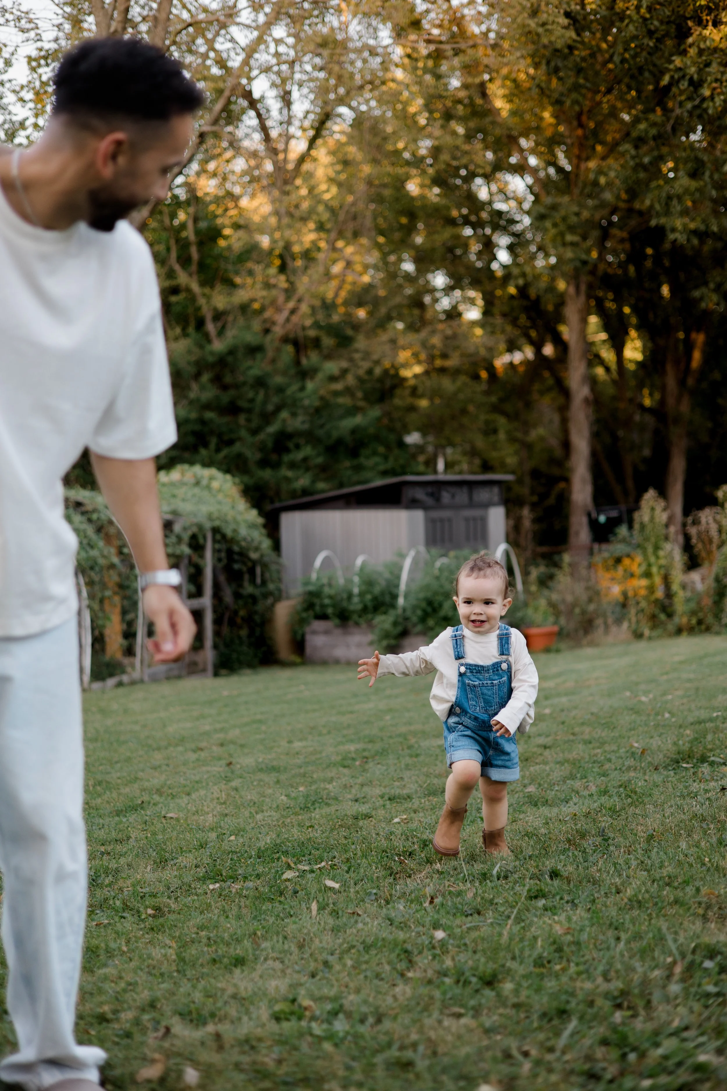 A young boy in denim overalls and boots running or playing outdoors on a grassy lawn, with a man partially visible on the left side and a shed or small building in the background surrounded by trees during the late afternoon or early evening.