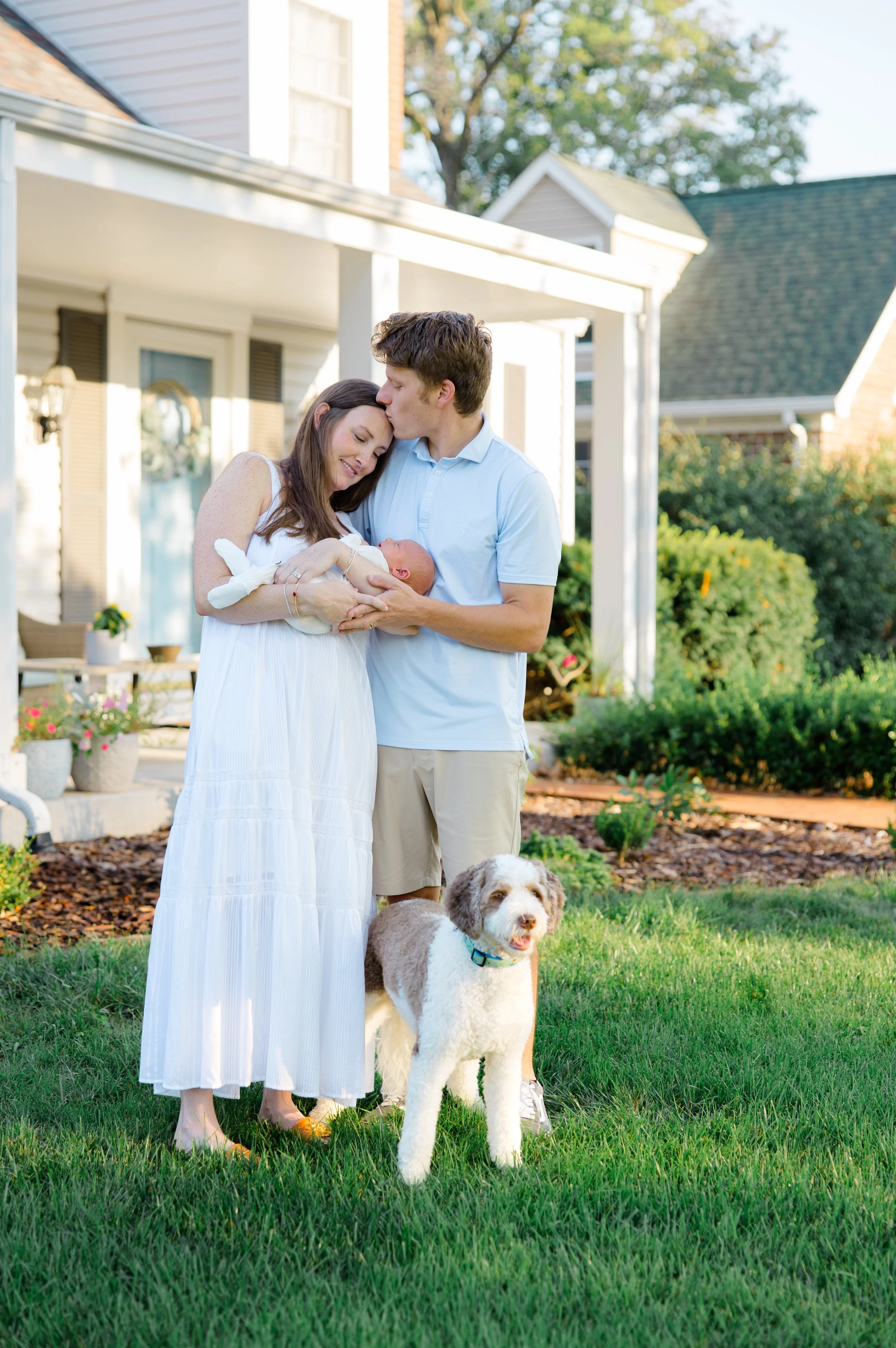A happy family of three with a dog in their yard, holding a newborn baby. The mother is wearing a white dress, the father is in a light blue shirt and khaki shorts, and the dog is a fluffy white and brown breed. The background features a house with a
