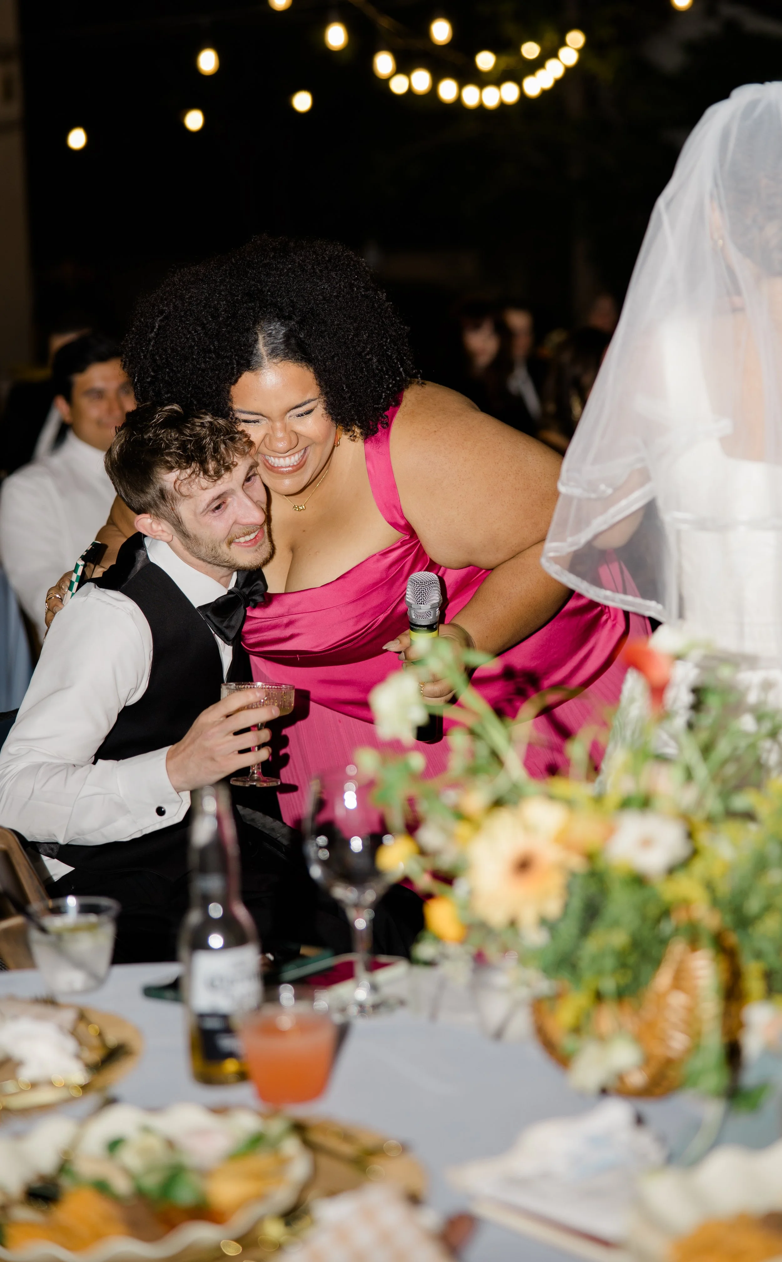 A bride in a white dress with a veil on her head kisses a woman in a bright pink dress, who is holding a microphone. The scene is at a wedding reception with string lights above and guests in the background. Table with floral centerpiece and drinks v