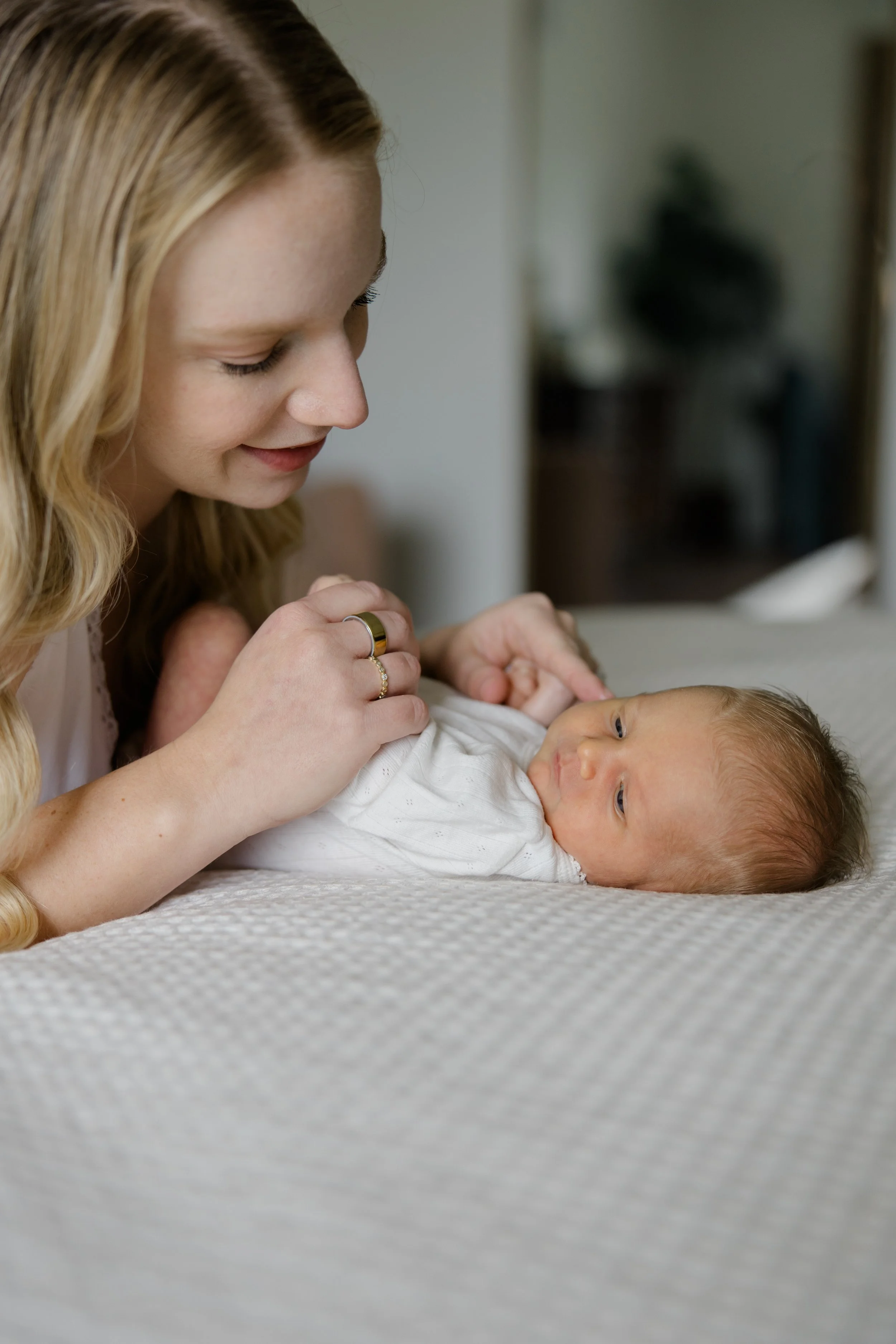 A young woman with long blonde hair smiling at a baby lying on a bed, with her hand resting on the baby's chest. The background is blurred.