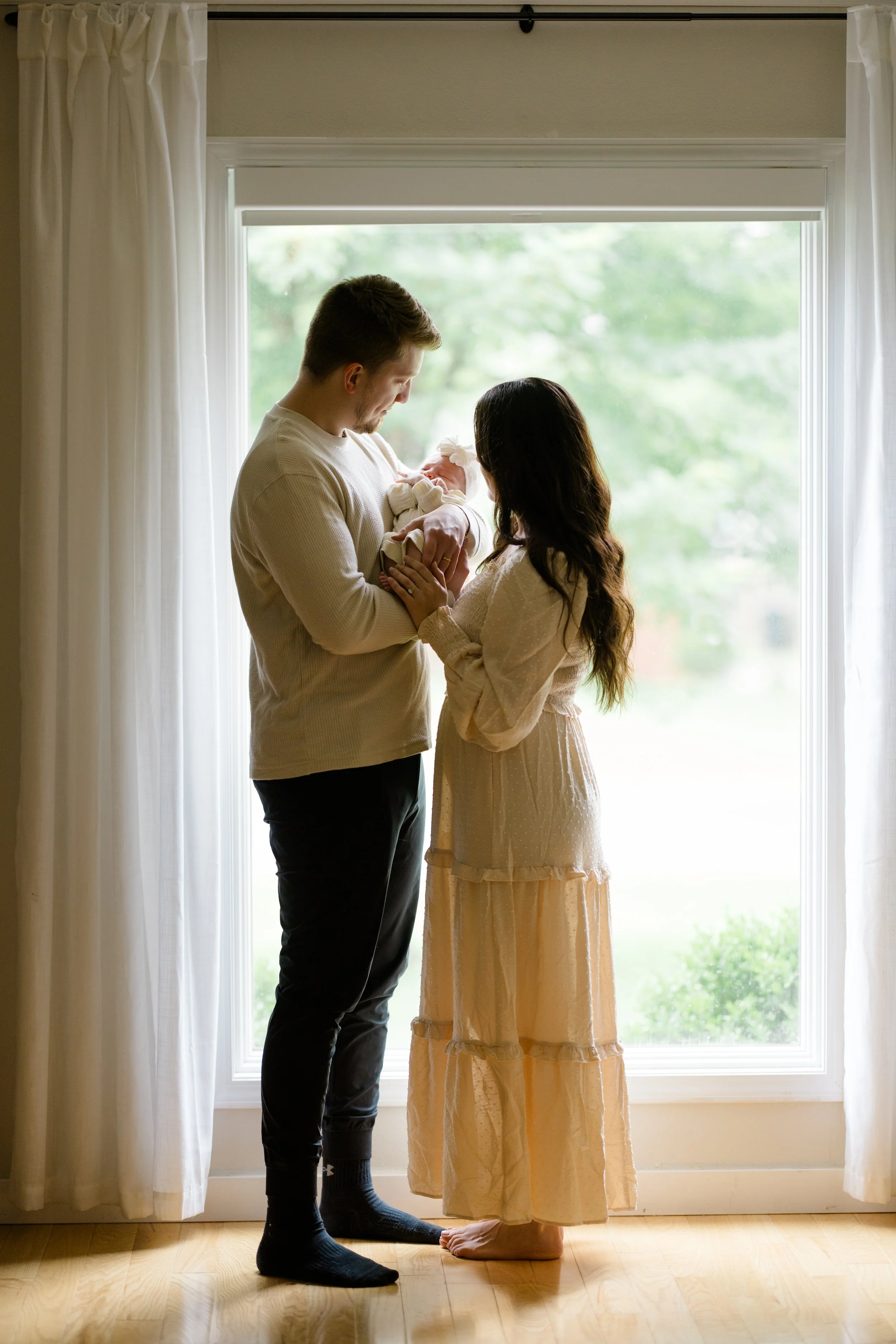 A man and woman standing in front of a large window, holding a newborn baby between them. The woman is looking down at the baby, and the man is looking at the baby, with the woman’s hands supporting the baby. The scene is bright with natural light, a