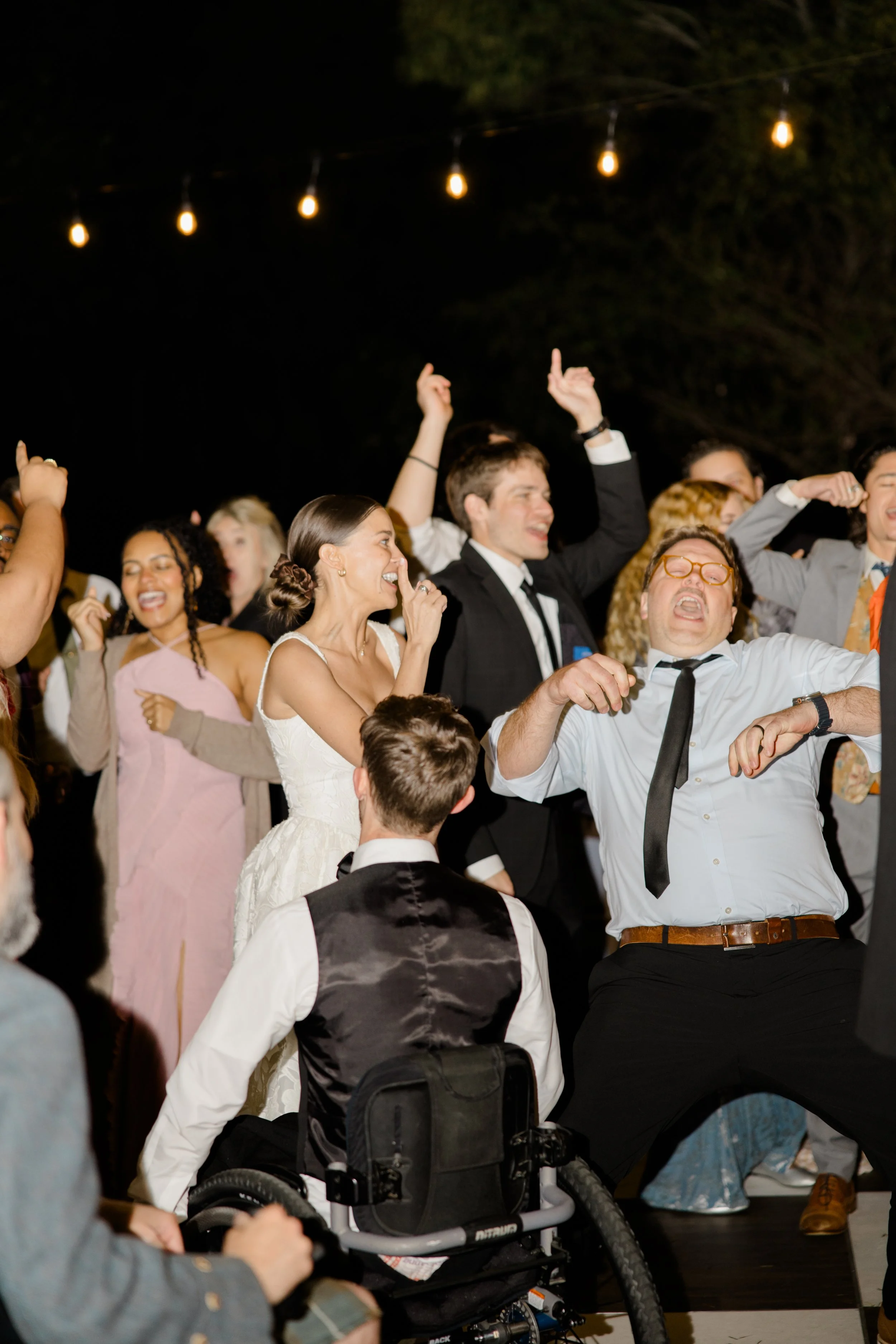 People dancing and celebrating at an outdoor event during night time, with string lights overhead.