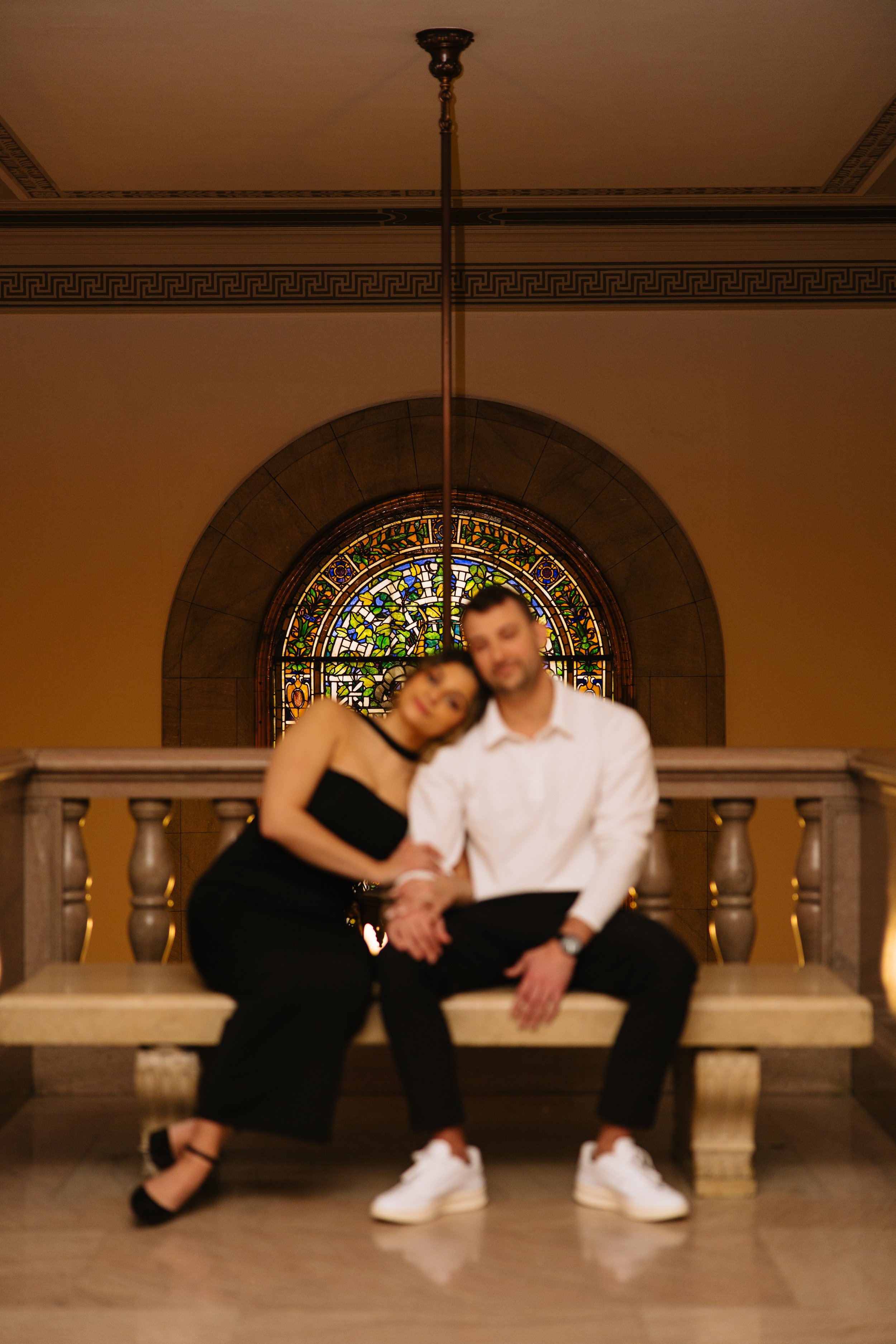 A couple sitting on steps in front of a colorful stained glass window in an elegant, historic building.