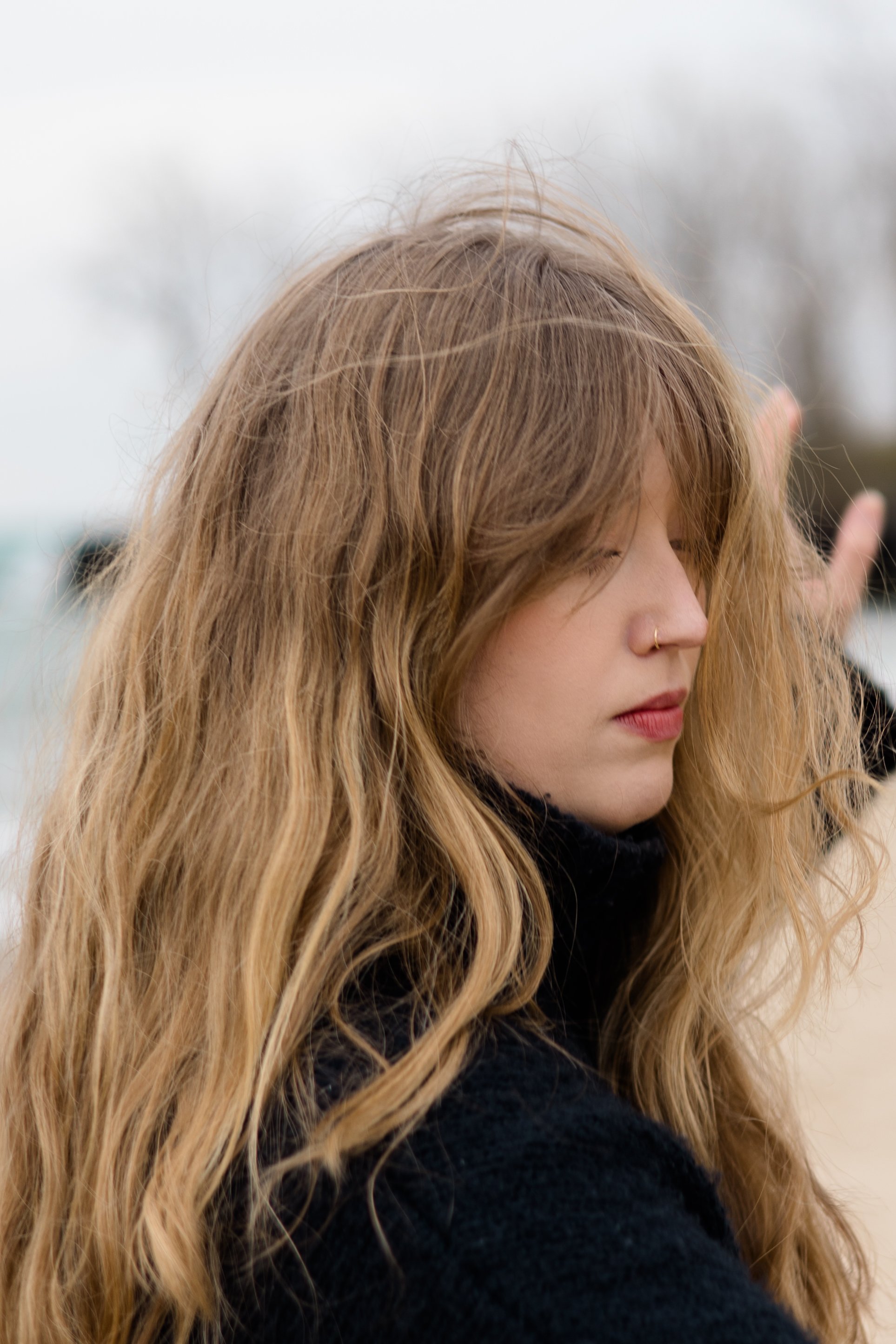 Close-up of a young woman with long, wavy blonde hair, wearing a black turtleneck, outdoors on a cloudy day, with trees in the blurred background.