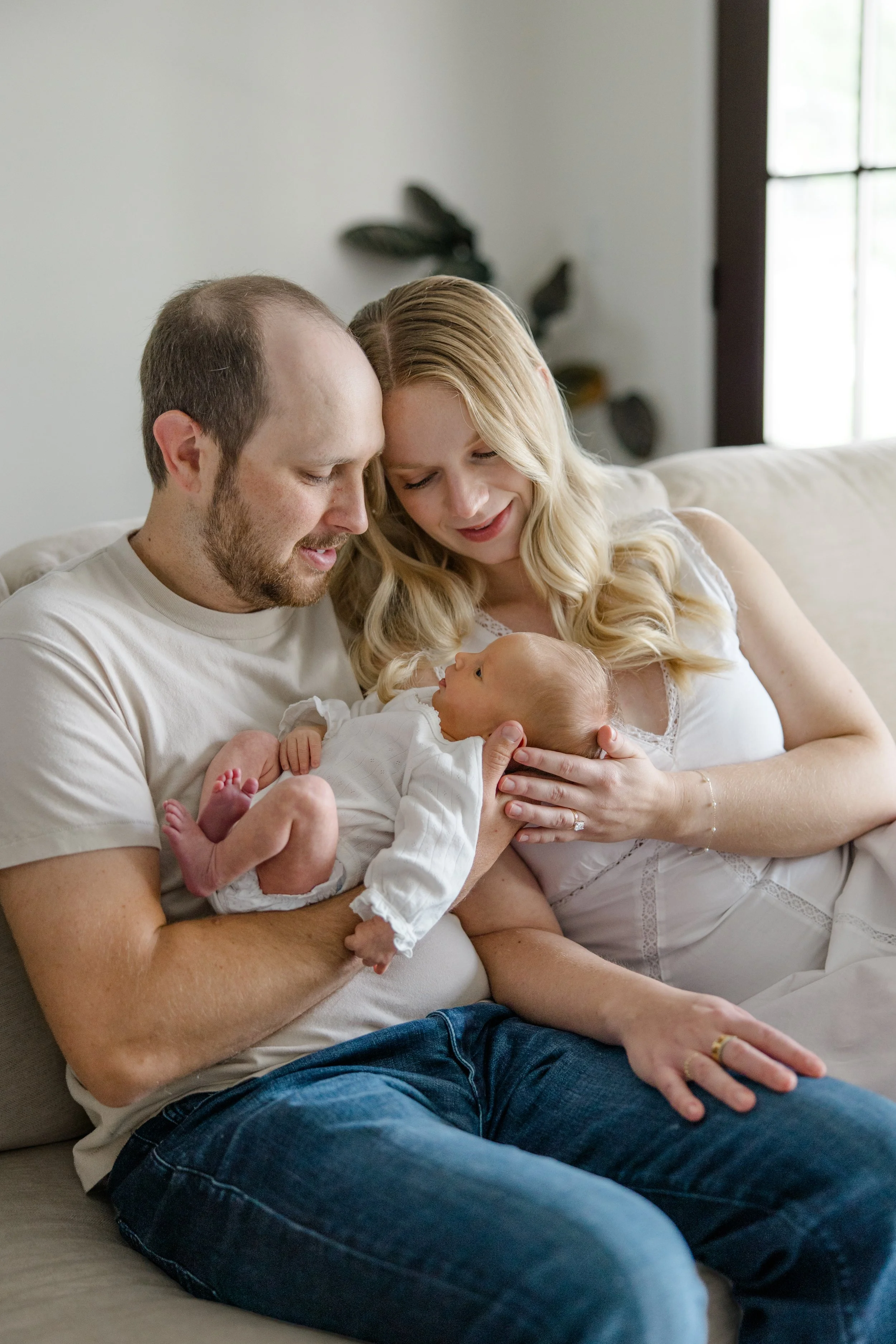 A family of three sitting on a couch, holding a newborn baby and smiling, in a cozy room with natural light.