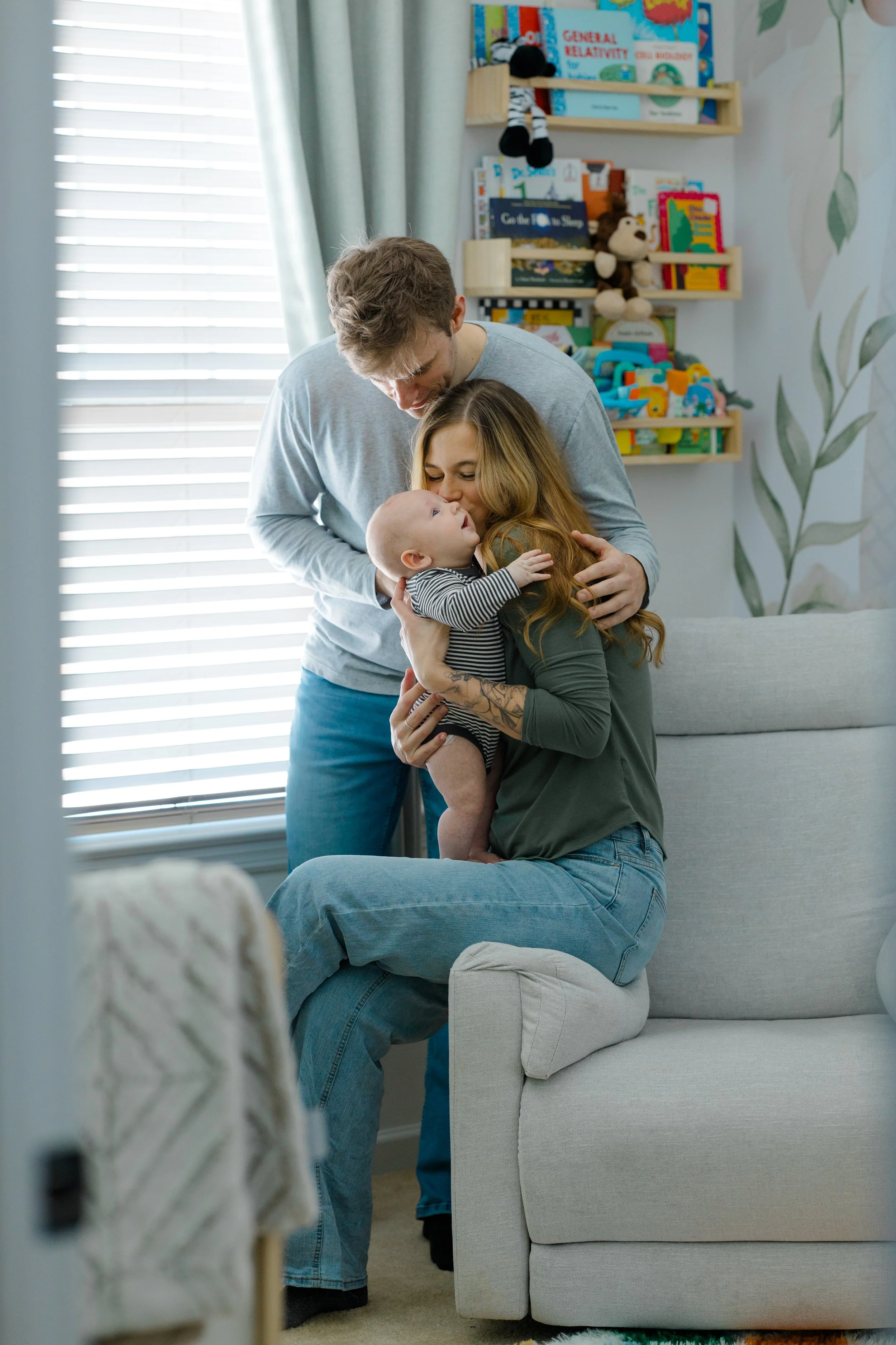 A woman is sitting on a couch holding a baby, while a man leans over them, and they are all touching foreheads. The background shows a bookshelf with toys and books, and a window with blinds.