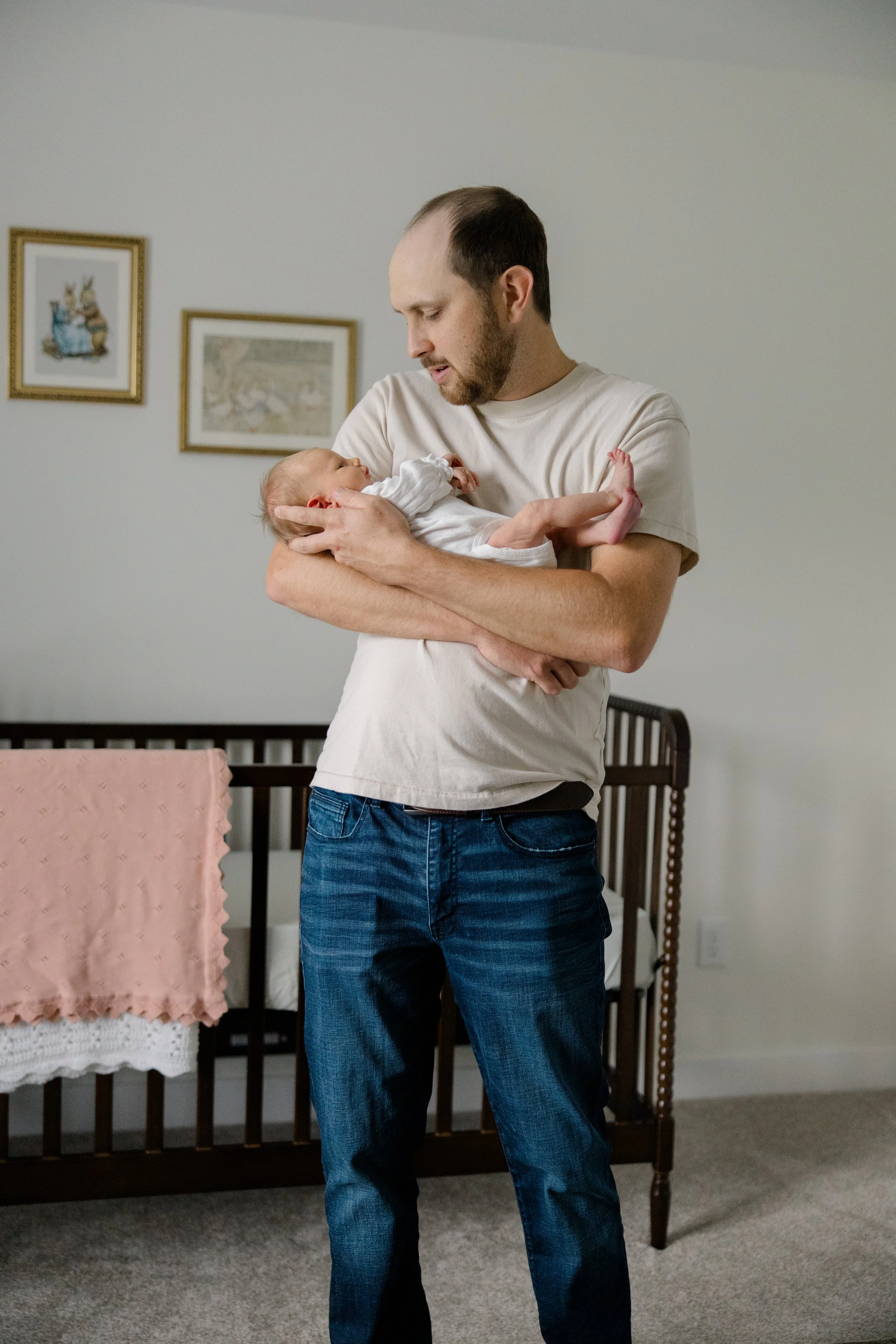 A man holding a baby in a bedroom with framed pictures on the wall and a crib with pink bedding.