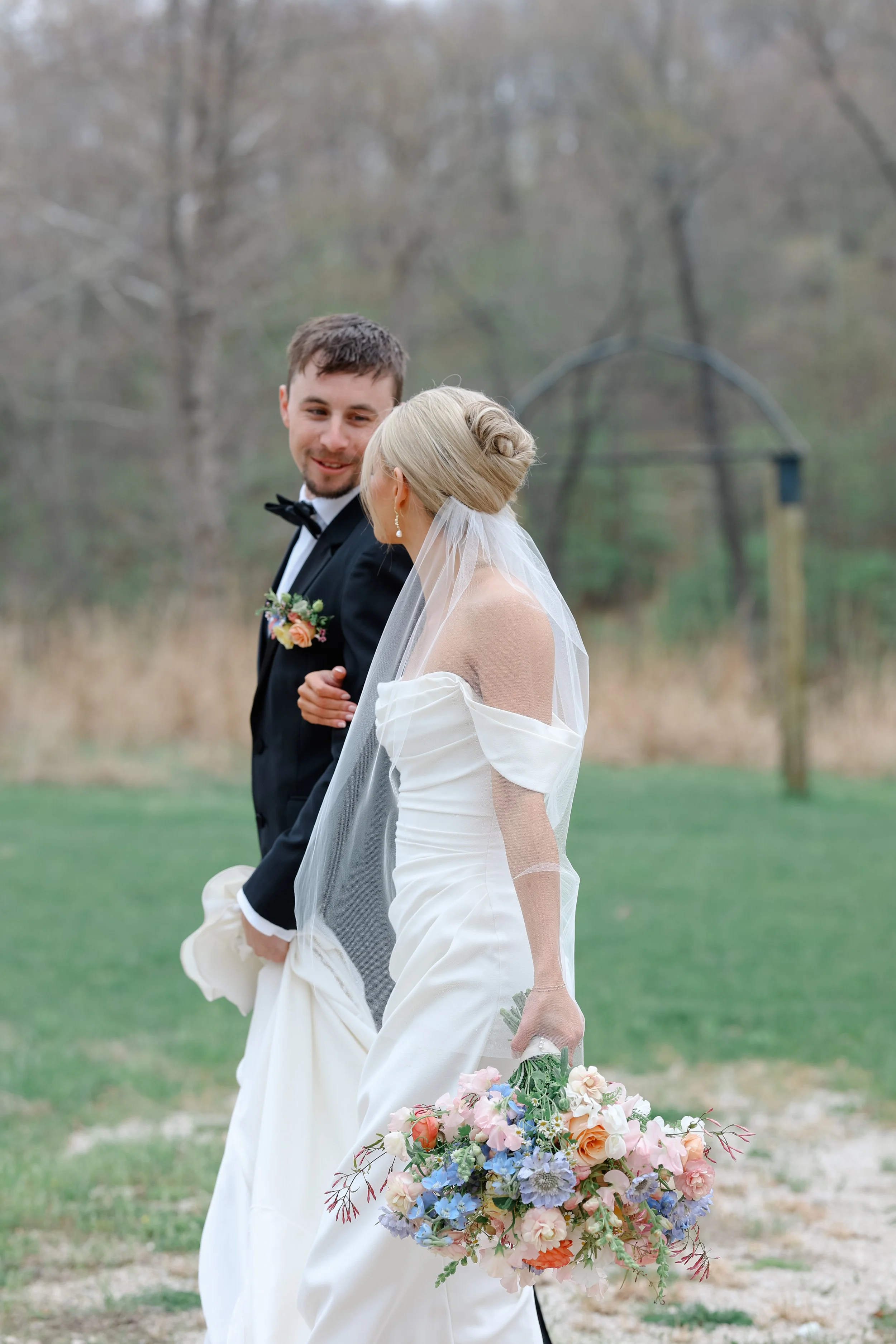 A bride and groom walking outside on their wedding day, holding hands and smiling at each other, with a bouquet of colorful flowers.