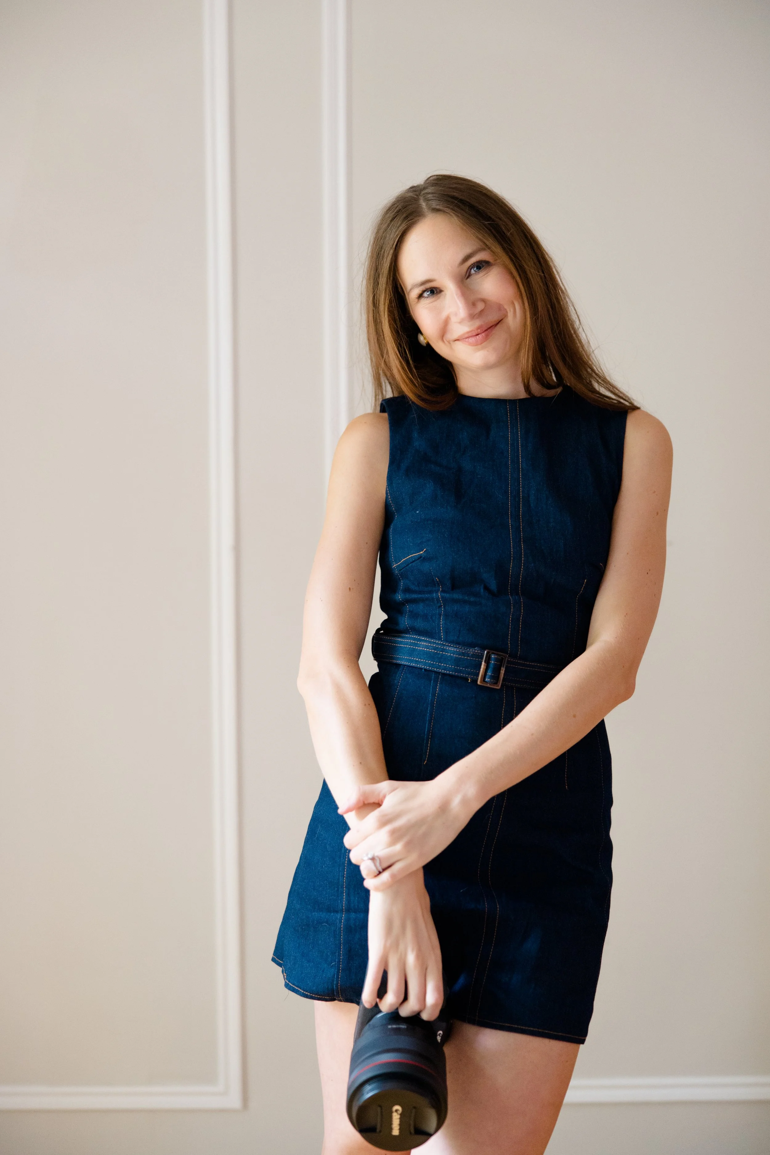 A young woman with long brown hair, wearing a sleeveless denim dress, smiling at the camera, holding a camera lens pointed at her thigh, standing against a plain cream-colored wall.