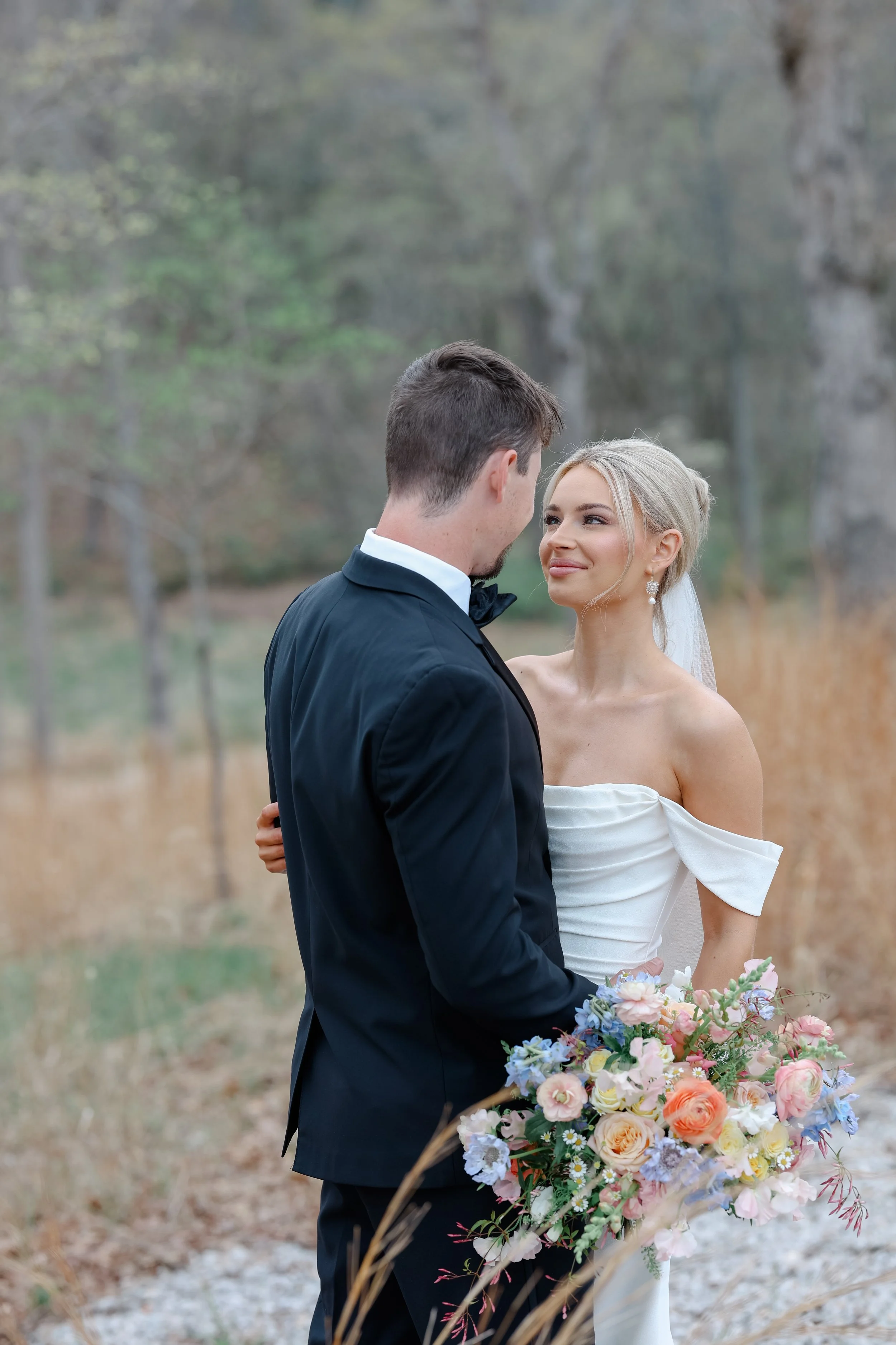 A newlywed couple gazing into each other's eyes during their outdoor wedding ceremony, surrounded by trees and holding a large bouquet of pastel-colored flowers.