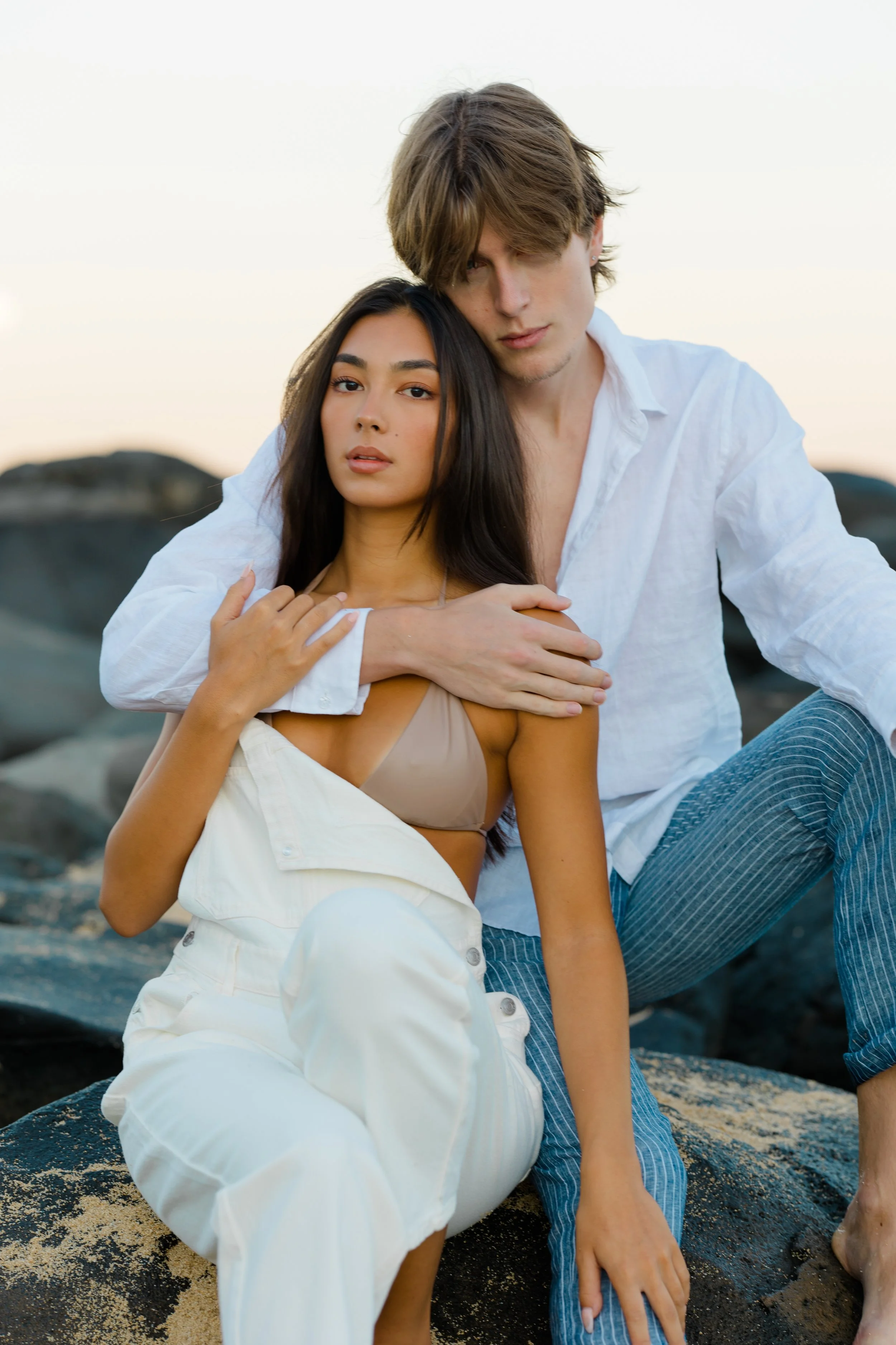 A young woman with long dark hair and a light beige top sitting on a rocky surface. A young man with shaggy light brown hair and a white shirt sitting behind her. The woman has her hand on her chest with a serious expression, while the man has his ar