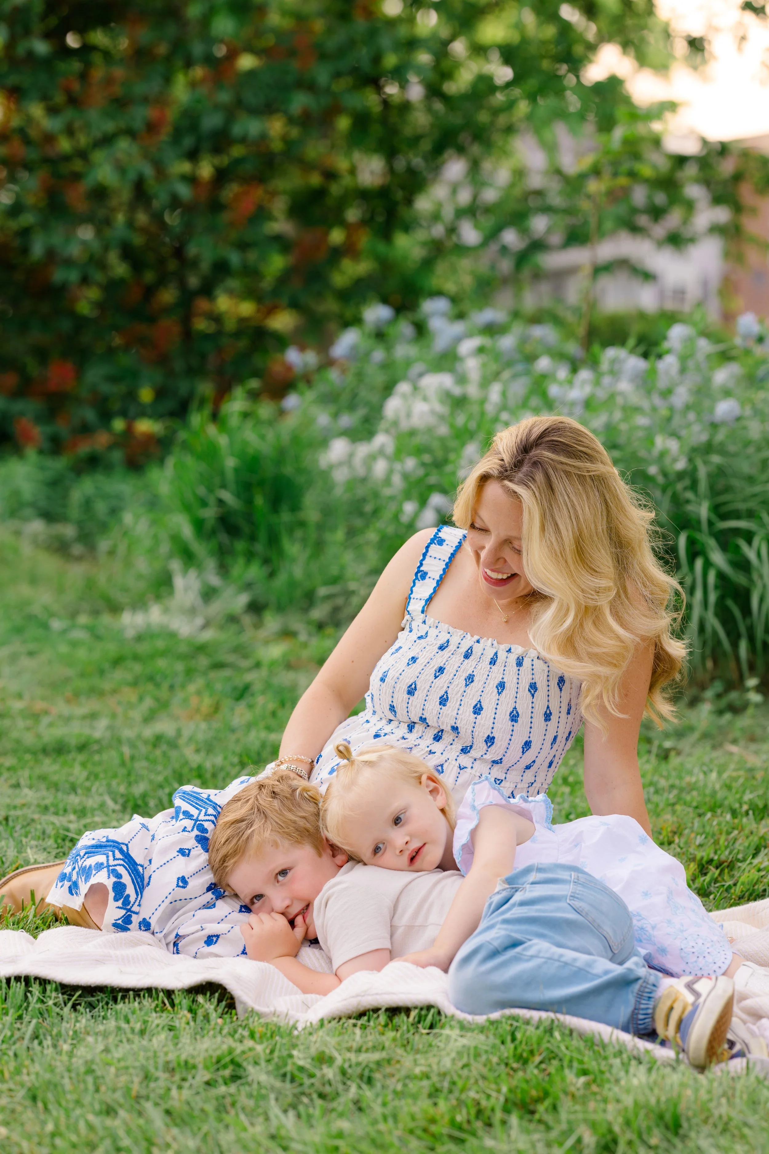 A woman with blonde hair smiling and sitting on the grass outdoors with three children, in a garden with greenery and white flowers in the background.