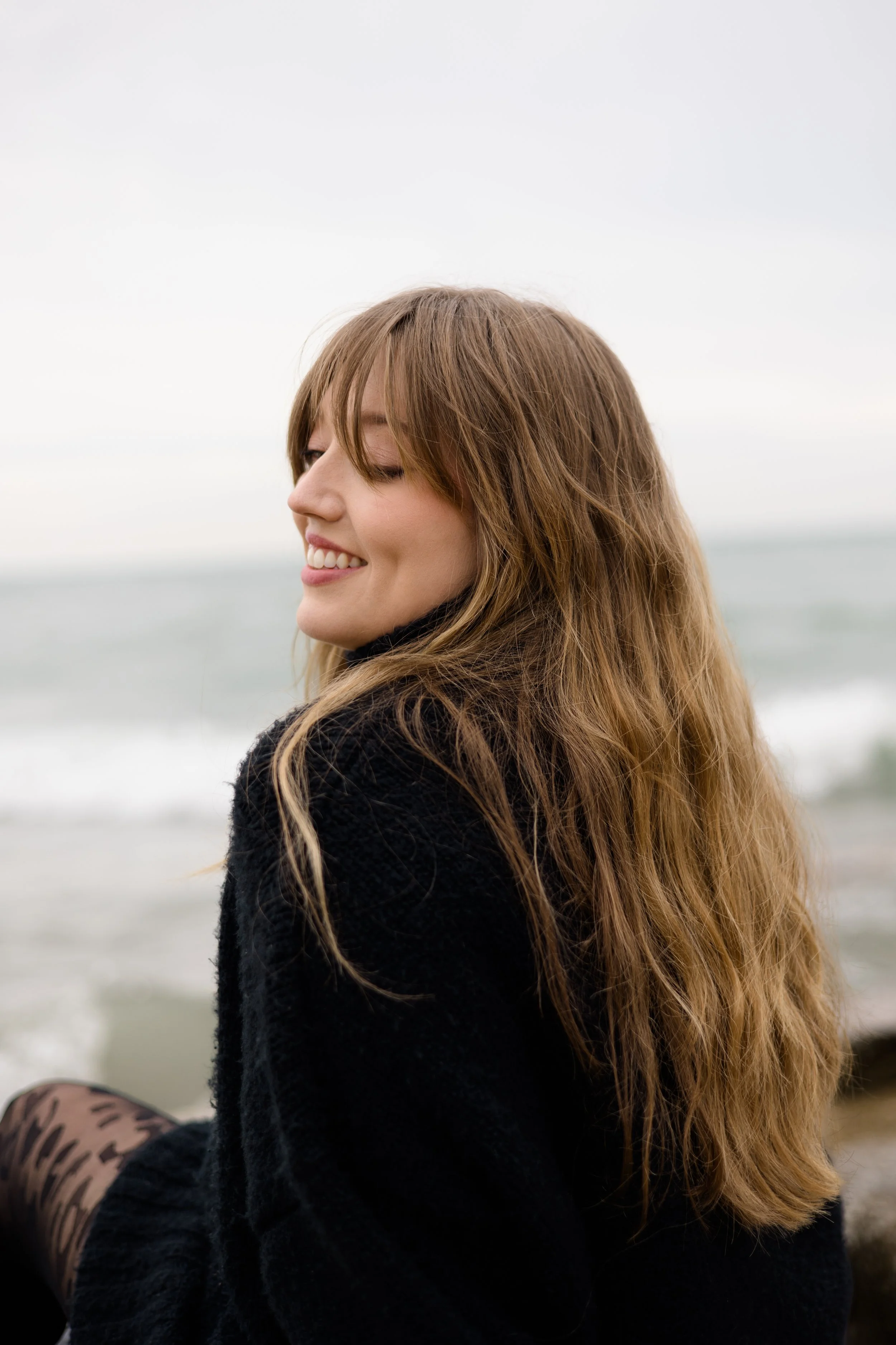 A smiling woman with long brown hair looking to her left, standing outdoors near water, wearing a black sweater and leopard print tights.