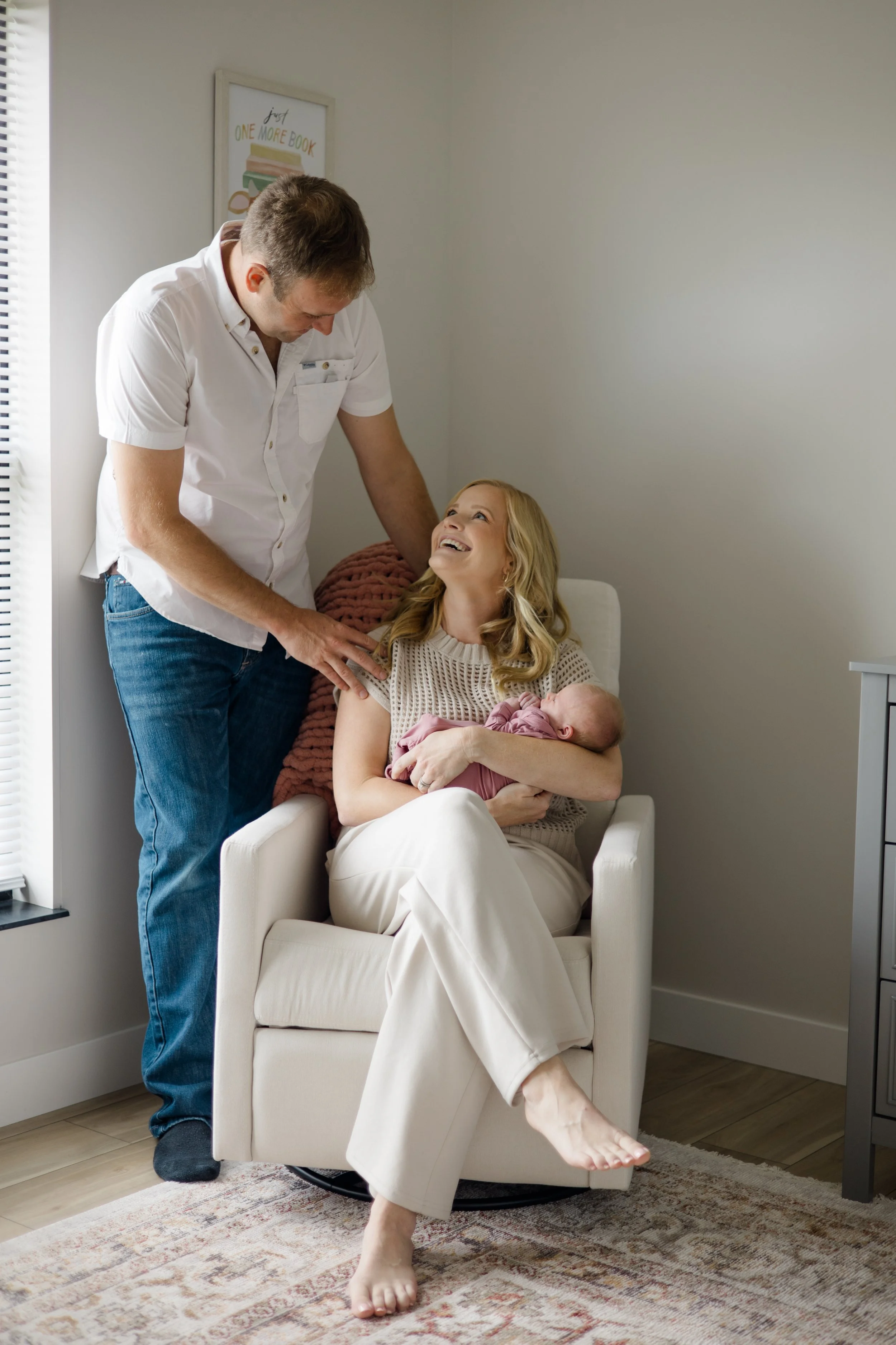 Woman sitting in a white armchair holding a newborn baby, smiling and looking up at a man who is leaning over her, with a woman in the background looking at the baby, in a home setting.