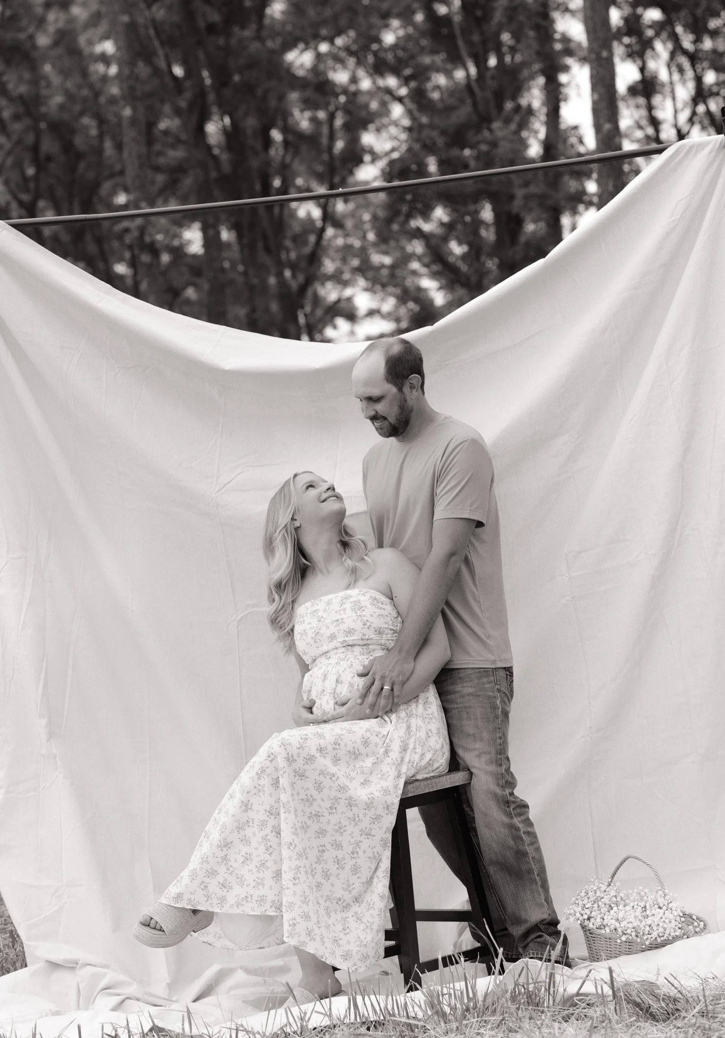 A black and white photo of a man and woman at an outdoor wedding photoshoot. The woman is sitting on a chair wearing a white dress, and the man is standing close to her. They are smiling and looking at each other. There is a white fabric backdrop han