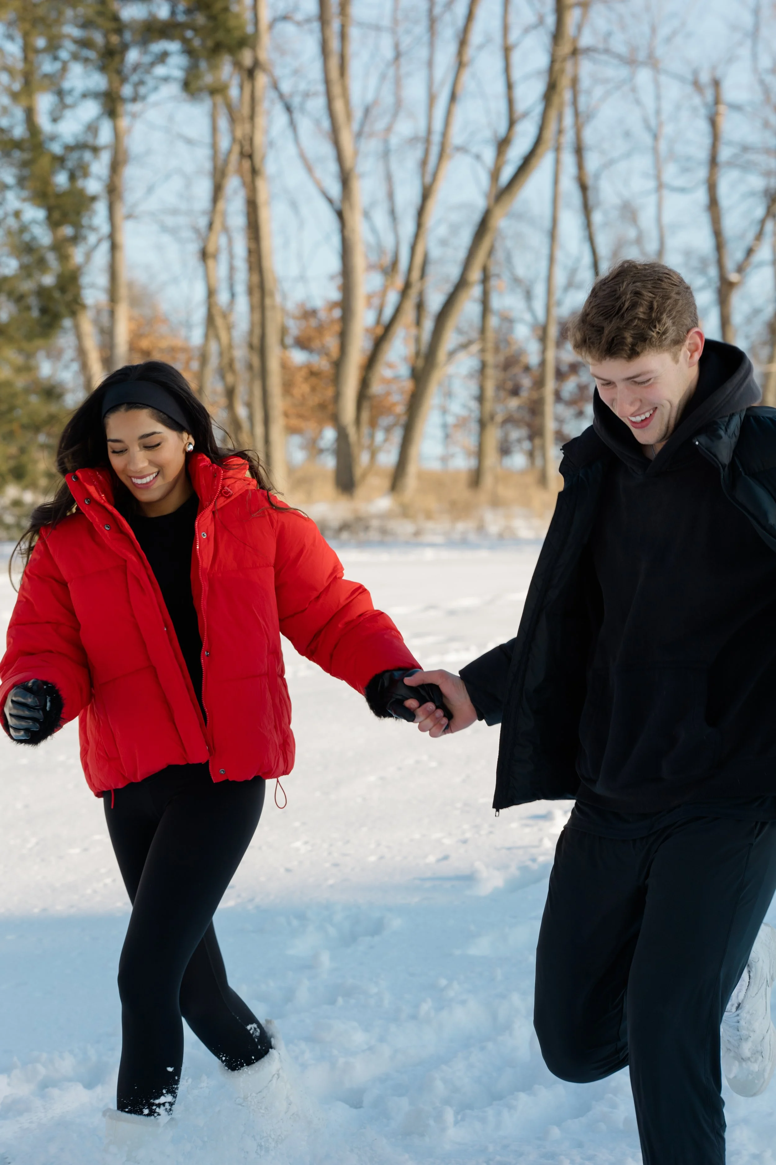 A young woman in a red winter jacket and black leggings holding hands with a young man in a black jacket, both smiling as they walk in the snow during winter.