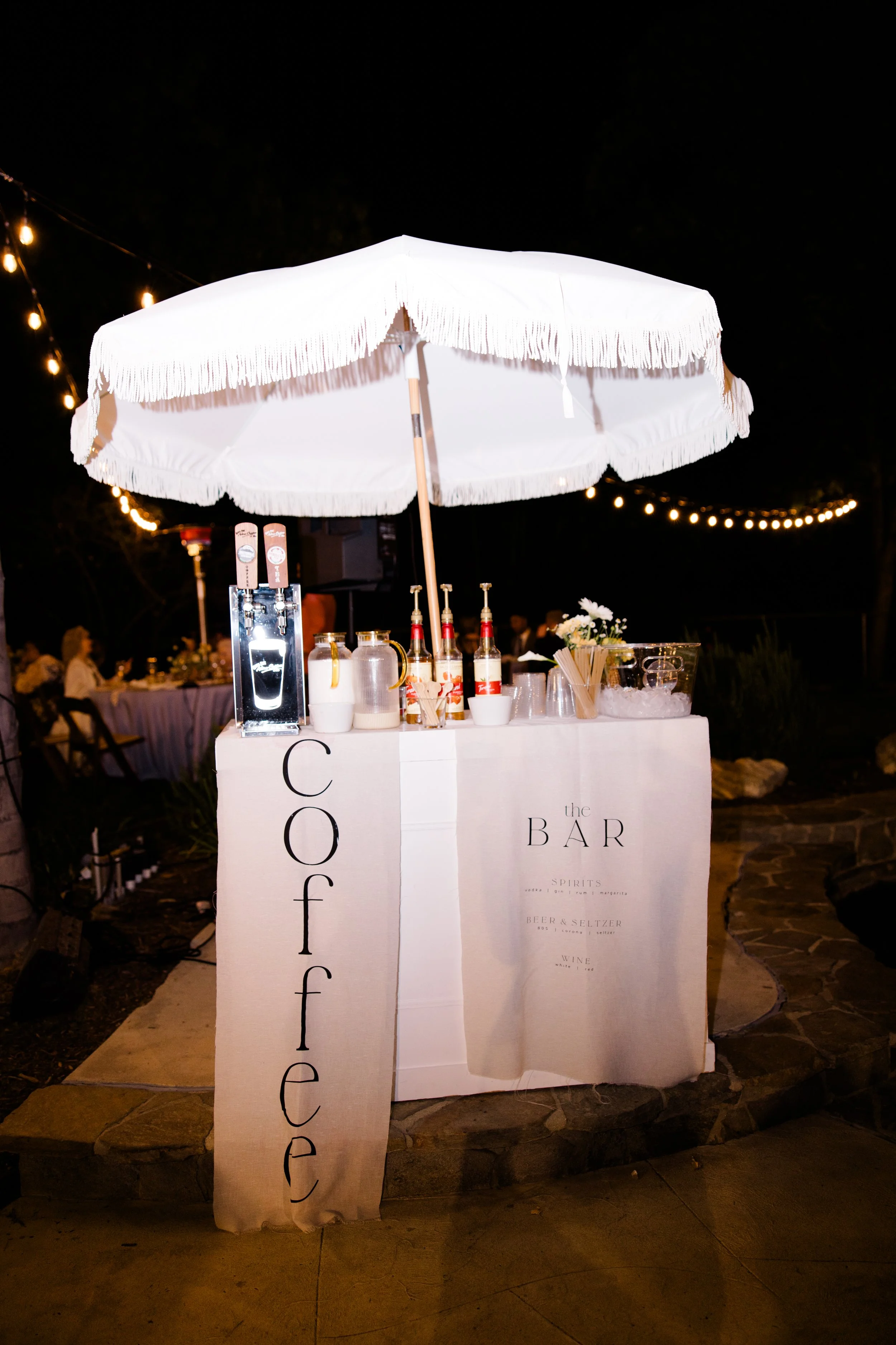 A nighttime scene of a white bar stand under a white fringed umbrella with bottled drinks, glasses, and a flower arrangement. String lights decorate the background.