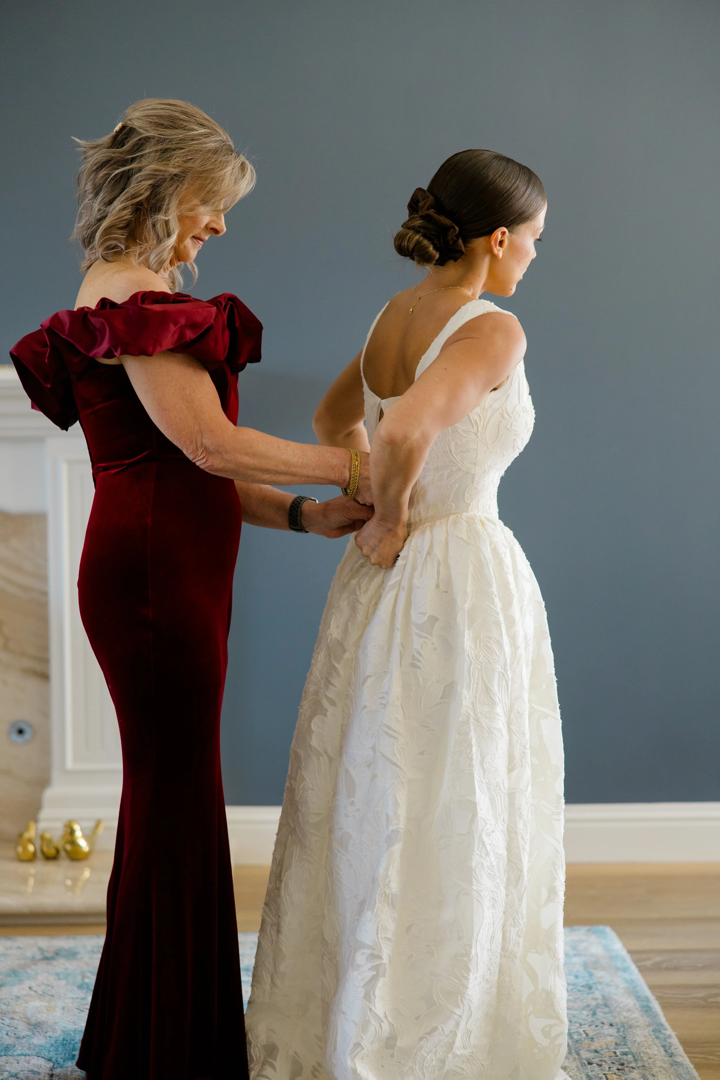 A woman in a white wedding dress standing with her back to a woman in a burgundy dress who is helping her zip up the dress in a room with a blue wall and a light-colored rug.