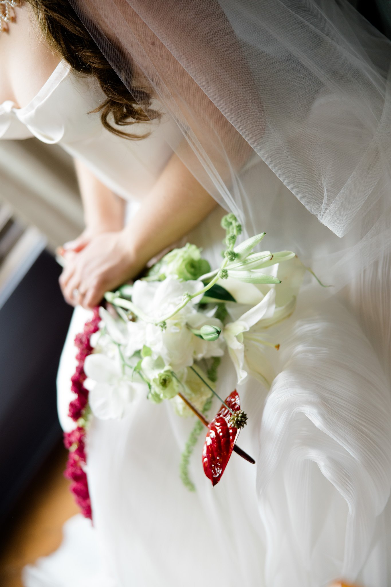 Close-up of a bride wearing a white dress, with a veil and intricate floral decorations on her dress, holding a bouquet of white flowers, with her arm resting on a surface.