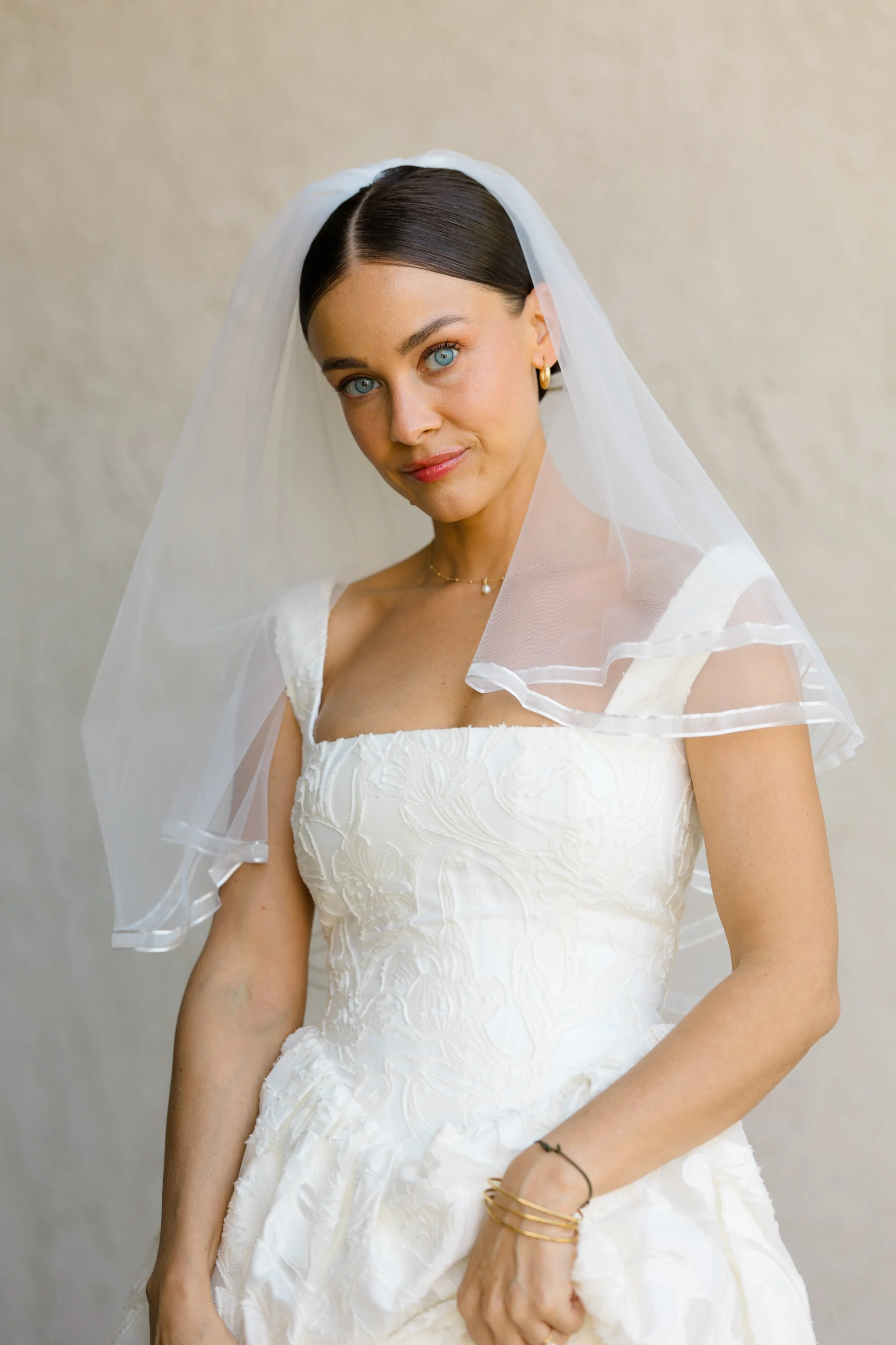 A woman in a wedding dress with lace details, wearing a veil, gold jewelry, and standing against a plain background.