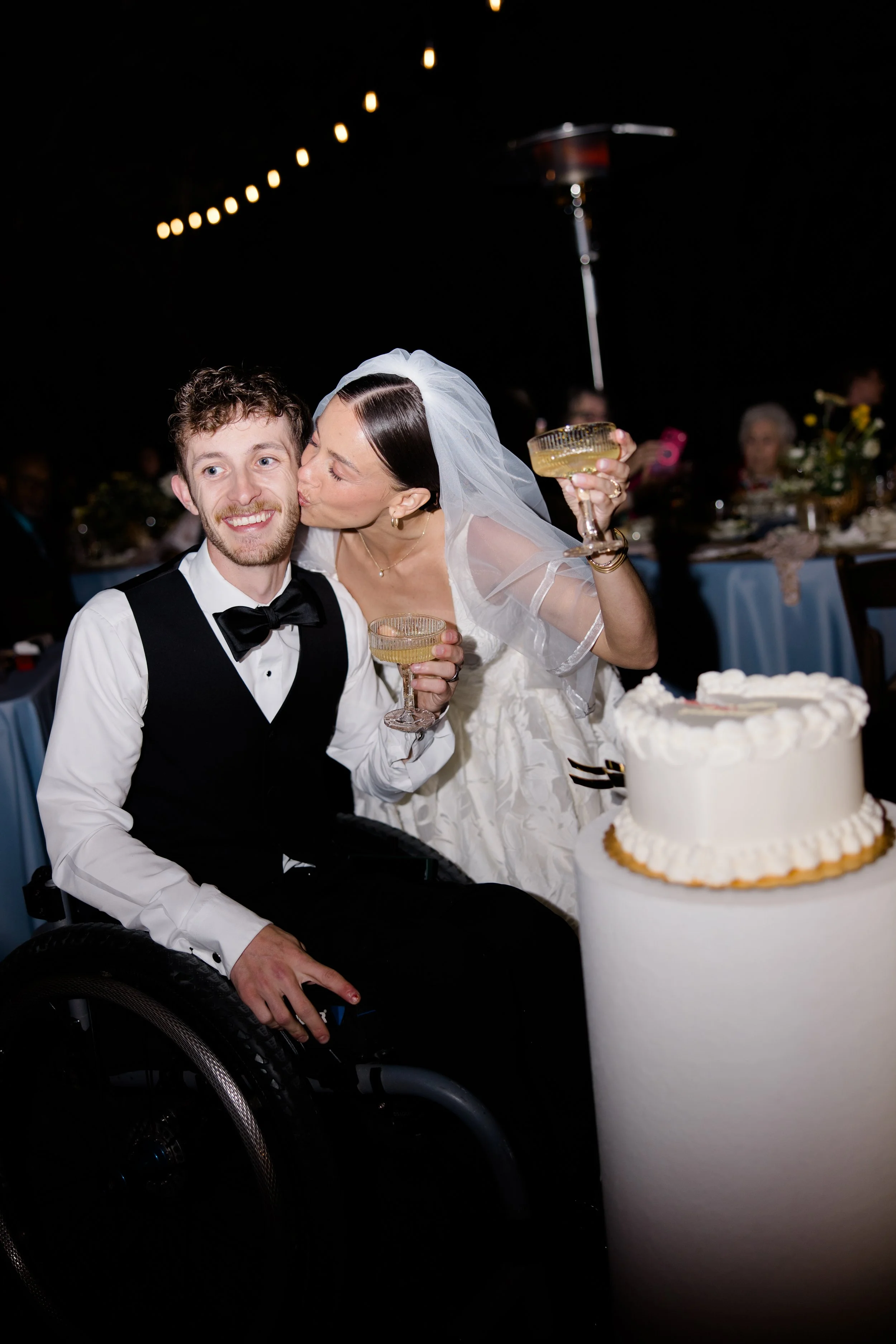 A bride leaning in to kiss a groom seated in a wheelchair, both holding drinks, at a wedding reception with a cake in the foreground and guests in the background. The bride wears a white veil and dress, and the groom wears a tuxedo with a bow tie.