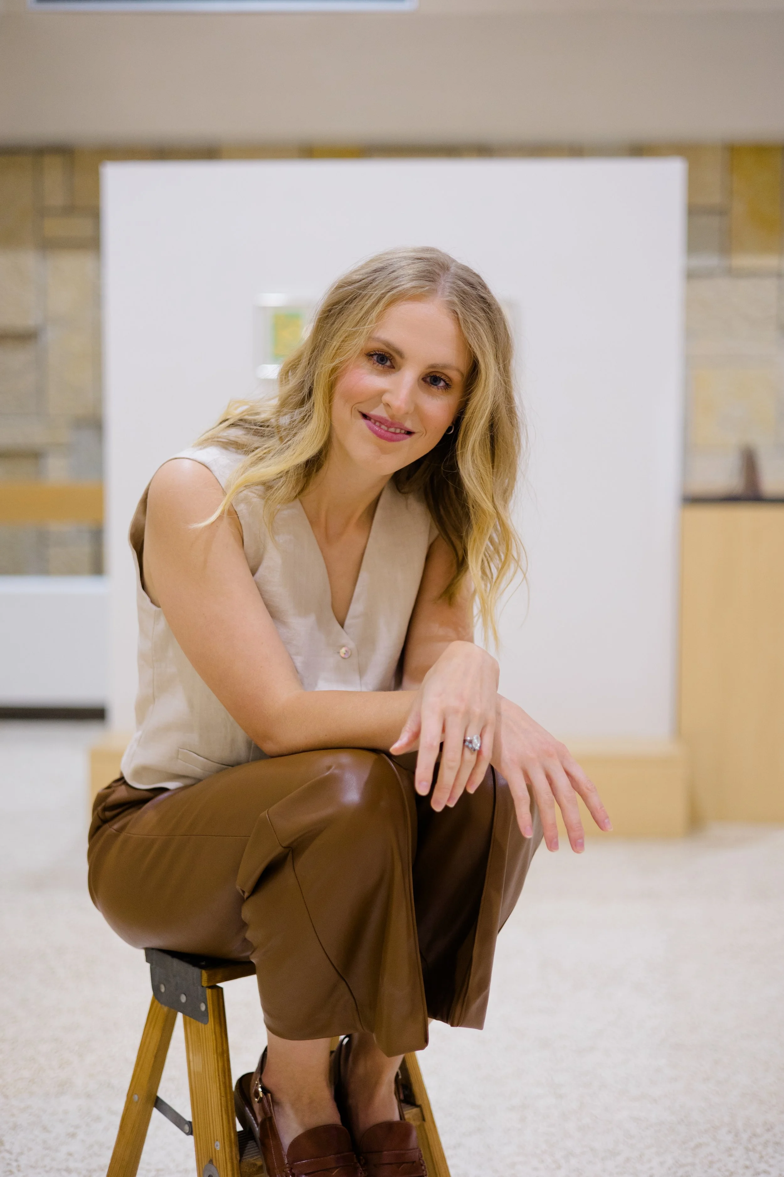 A woman with long blonde hair, wearing a sleeveless beige top and brown leather pants, sitting on a wooden stool with her hands resting on her knees, smiling at the camera in a room with a neutral-colored wall and a small piece of artwork in the back