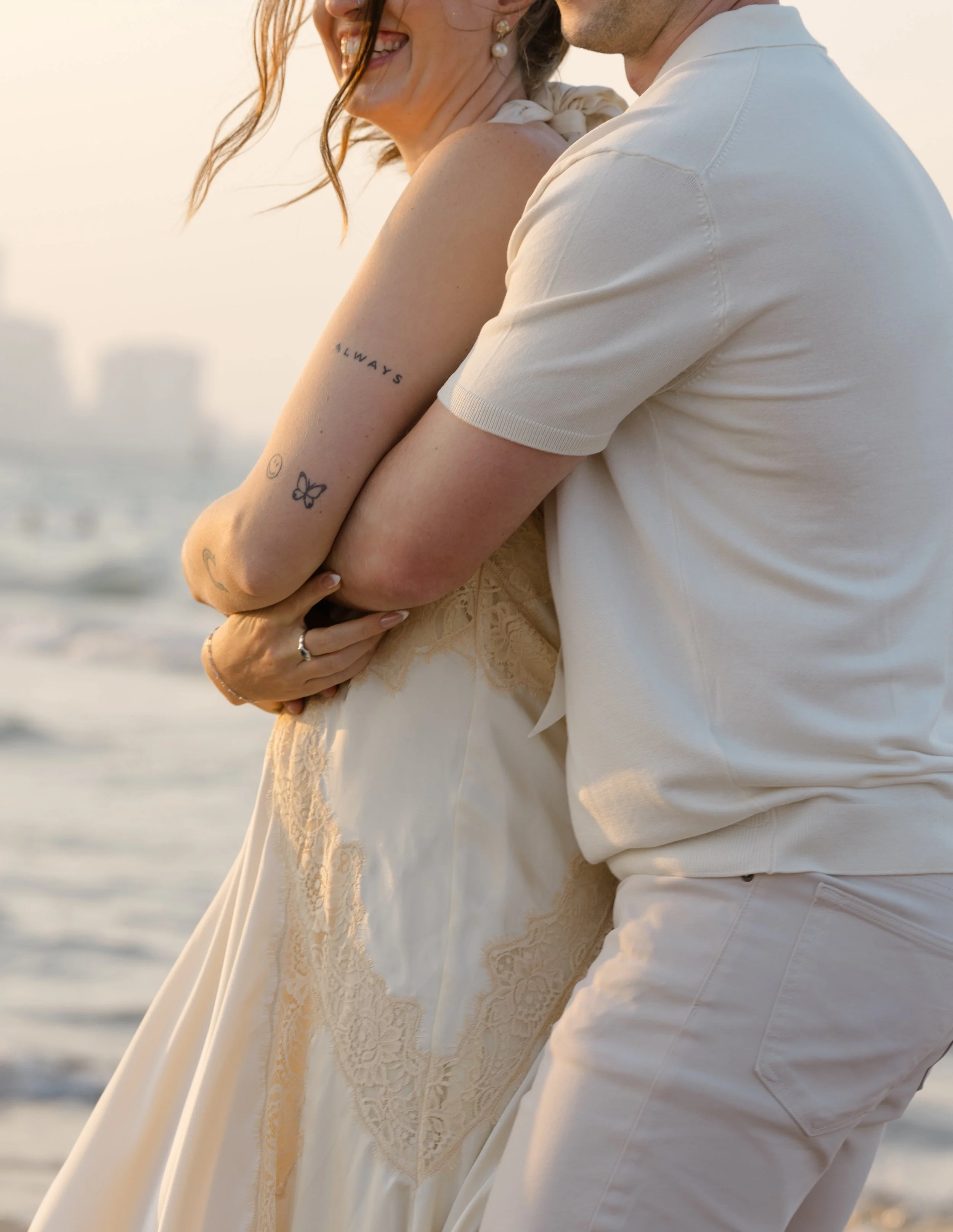 A couple on the beach, smiling and hugging, with the sunset reflecting on the water in the background.