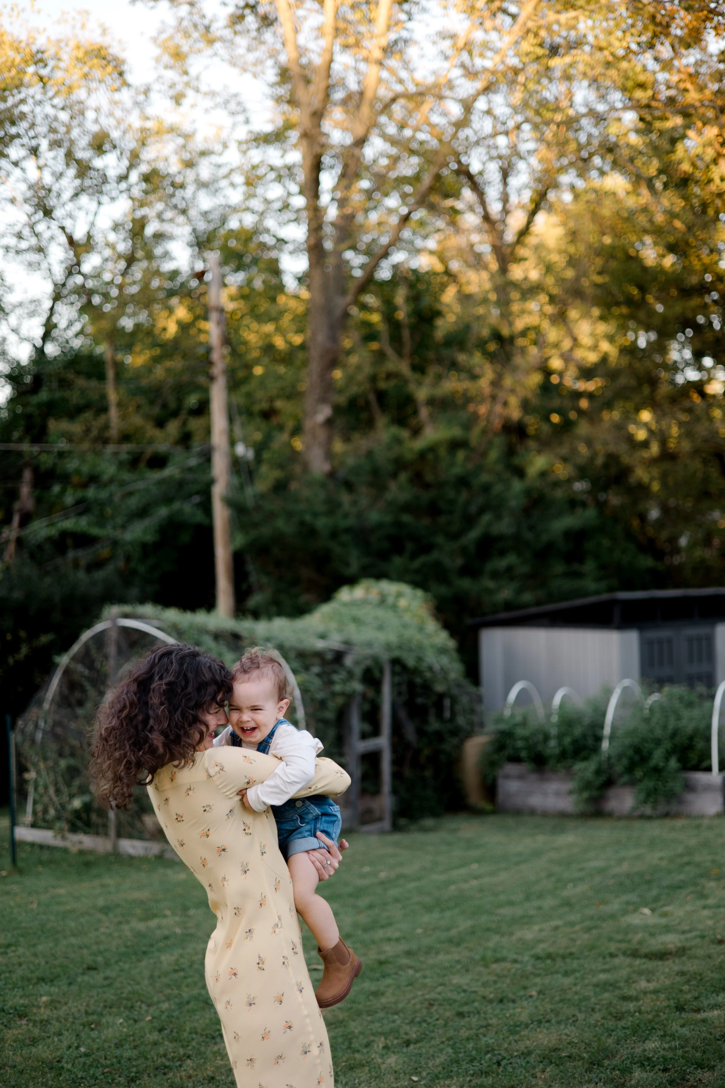 A woman with curly dark hair lifting a young boy with light hair and wearing a white shirt and denim shorts, both smiling and enjoying their time outdoors in a backyard with green grass, trees, and garden beds.