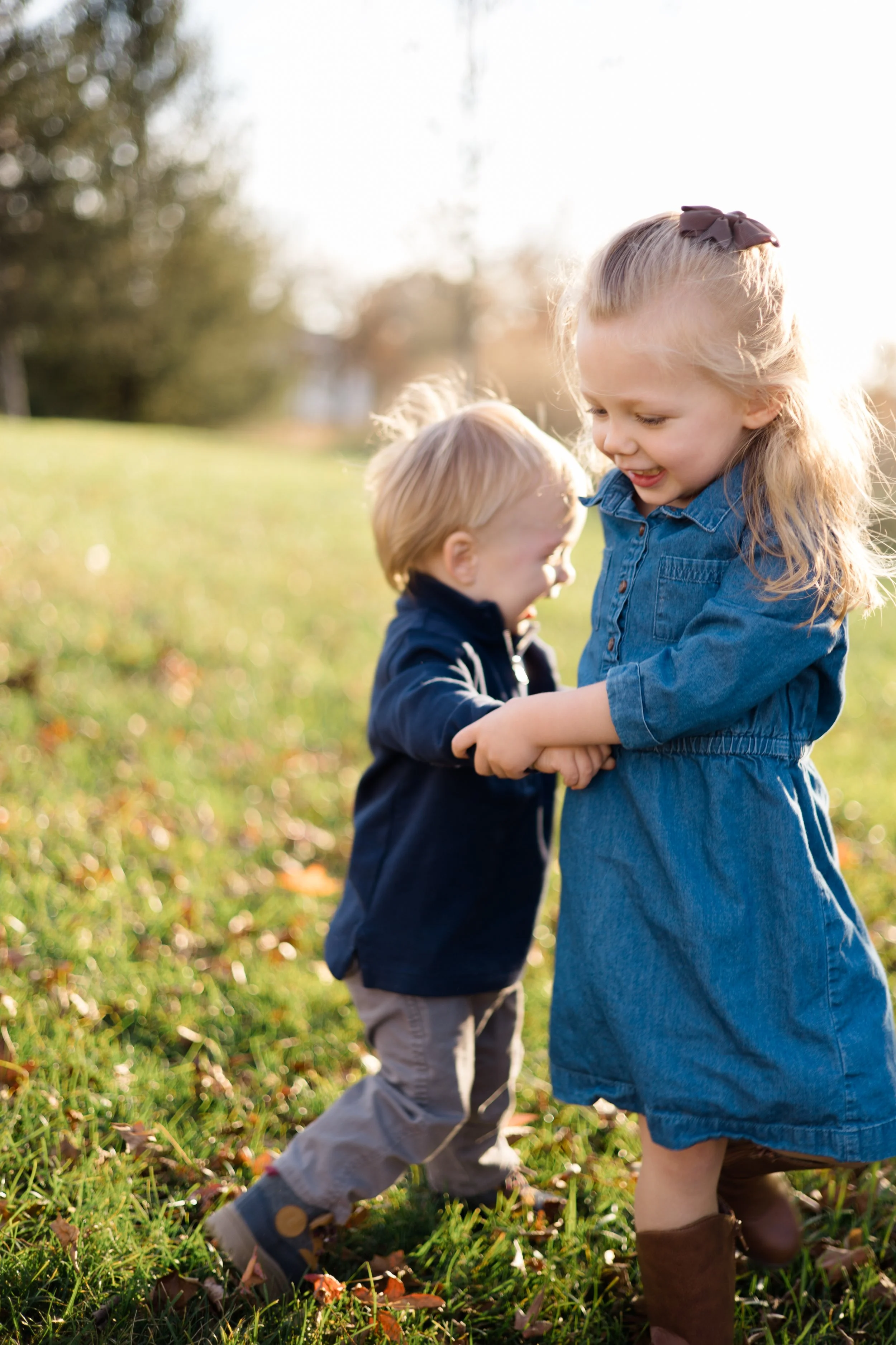 Two young children, a girl and a boy, playing together outdoors in a grassy area on a sunny day, holding hands and smiling.