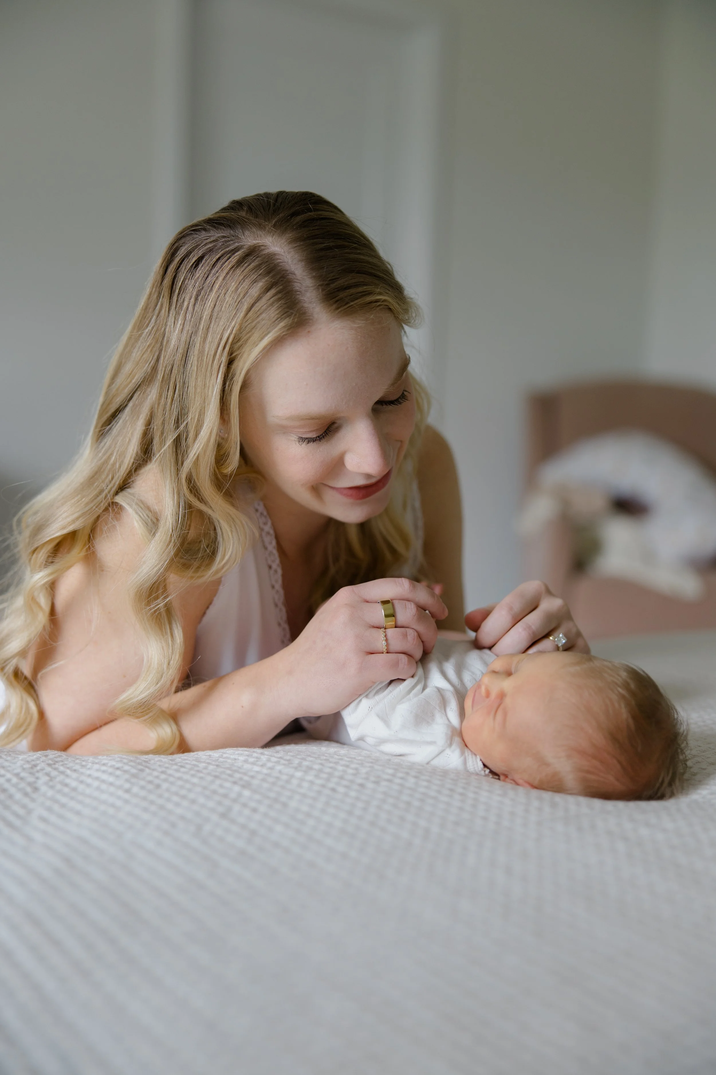 A woman with long blond hair with soft curls, smiling while leaning over a newborn baby lying on a bed. The woman is wearing a gold ring and a small ring on each hand. The background shows a blurred bed with a wooden headboard.